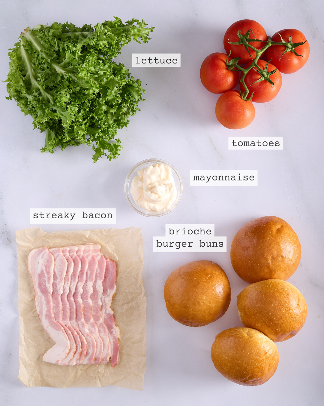 Flat lay of ingredients for Korean-style pork burger assembly including brioche burger buns, streaky bacon, lettuce, vine tomatoes and mayonnaise, each clearly labelled on a white marble surface.