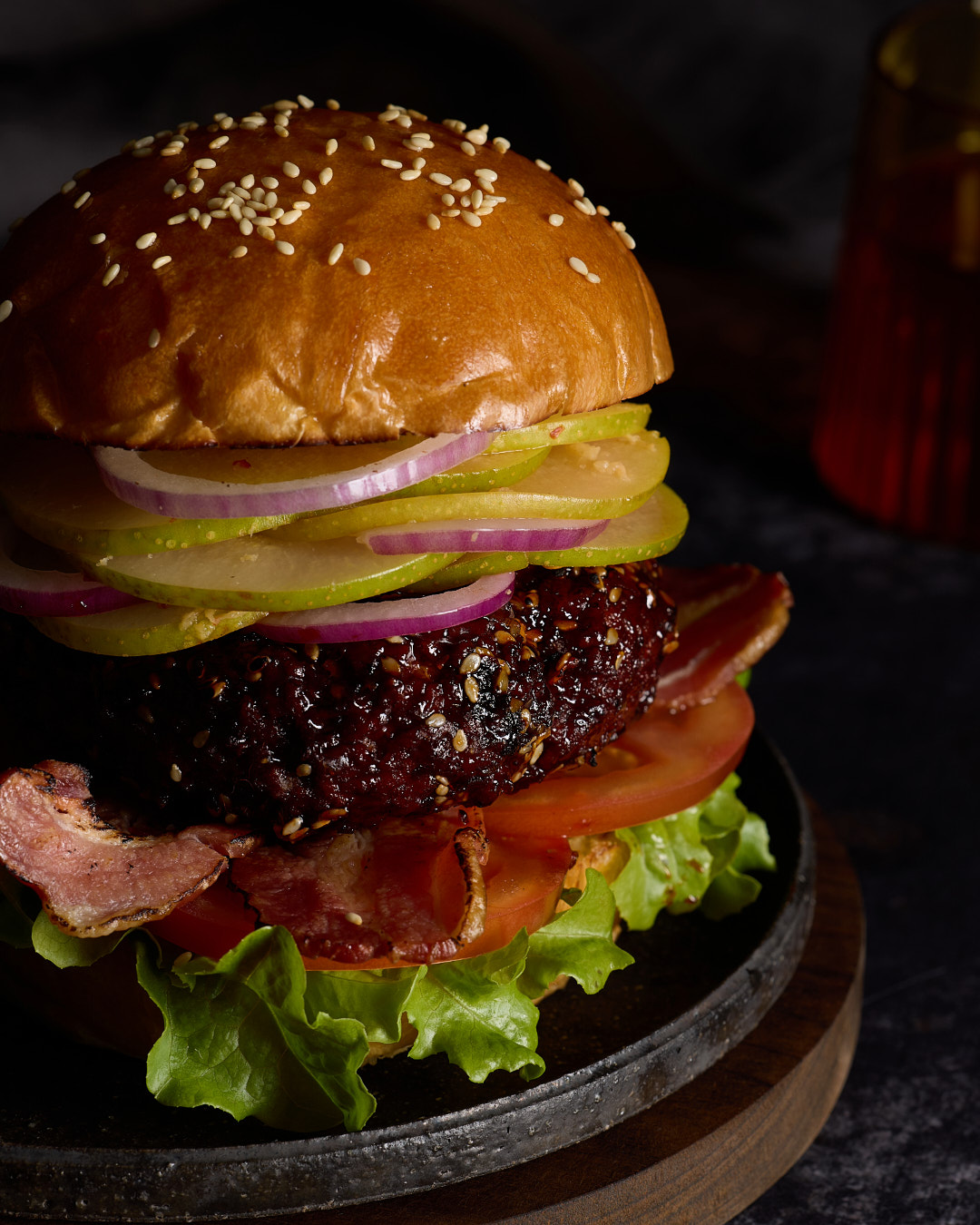 Korean-style pork burger with gochujang glazed pork patty, fresh nashi kimchi, red onion, streaky bacon, lettuce and tomato on a brioche bun with sesame seeds, stacked high on a dark wooden board against a dark moody background.