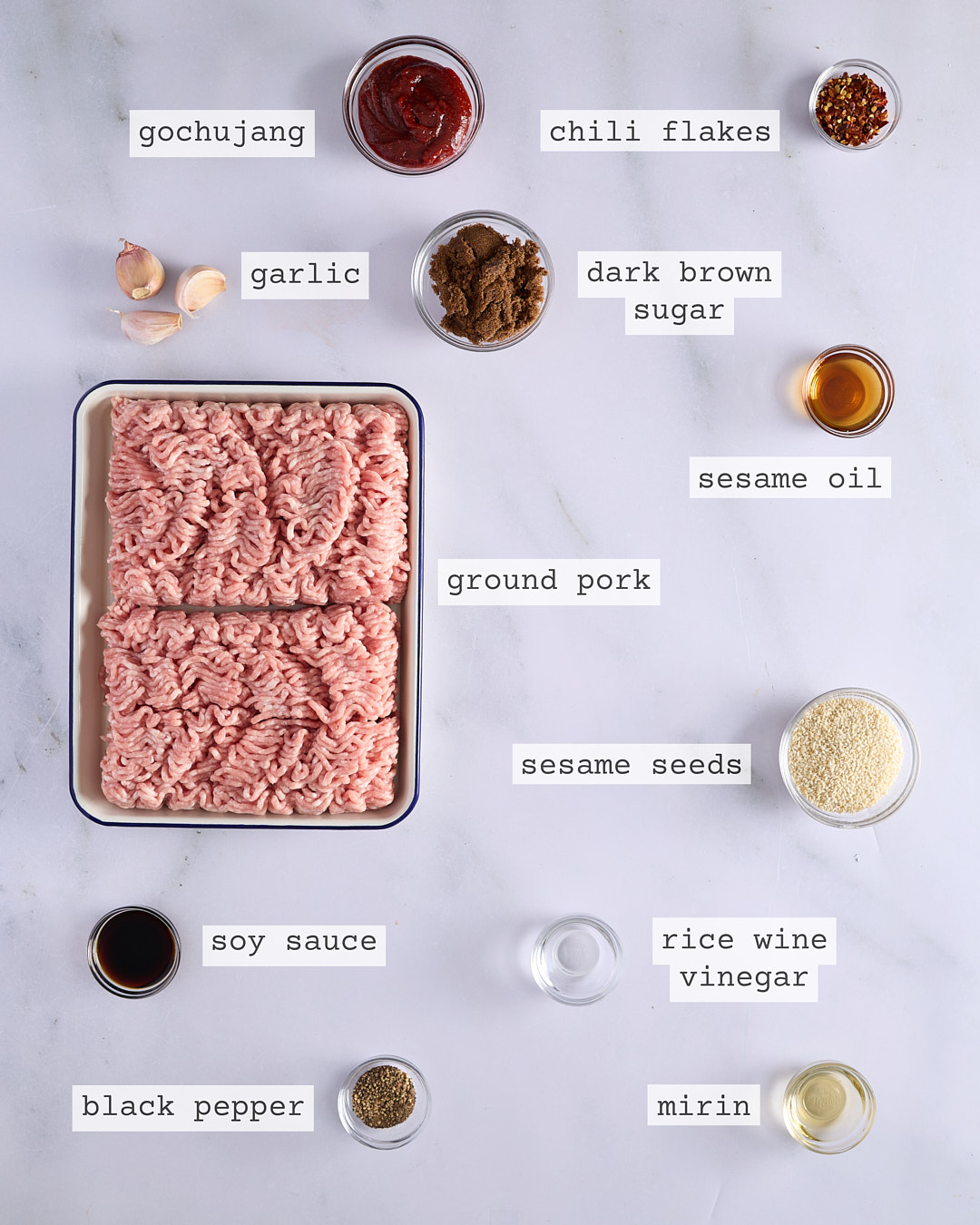 Flat lay of ingredients for gochujang pork burger including pork mince, gochujang, chilli flakes, garlic, dark brown sugar, sesame oil, sesame seeds, soy sauce, rice wine vinegar, mirin and black pepper, each clearly labelled on a white marble surface.