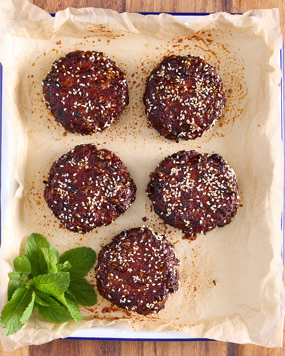 5 gochujang glazed pork burger patties topped with toasted sesame seeds on a baking paper lined enamel tray, deeply lacquered and cooked through, with fresh mint alongside.