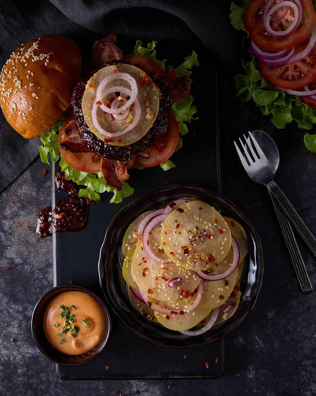 Korean-style pork burger with gochujang glazed pork patty, streaky bacon, lettuce, tomato, fresh nashi kimchi and spicy mayo on a brioche bun, served open on a dark board alongside a bowl of fresh nashi kimchi with red onion, chilli flakes and sesame seeds, and a small bowl of gochujang mayo, on a dark moody surface with fork and spoon alongside.