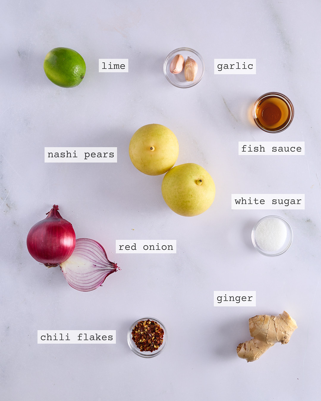 Flat lay of ingredients for fresh nashi kimchi including nashi pears, red onion, chilli flakes, garlic, ginger, fish sauce, white sugar and lime, each clearly labelled on a white marble surface.