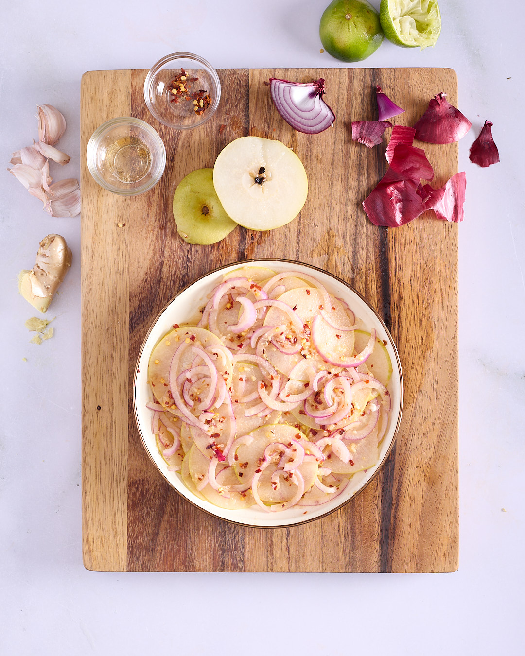 Fresh nashi kimchi with paper-thin nashi pear and red onion slices dressed in kimchi seasoning in a wide bowl on a wooden board, with halved nashi pear, red onion, garlic, ginger, chilli flakes and lime alongside.