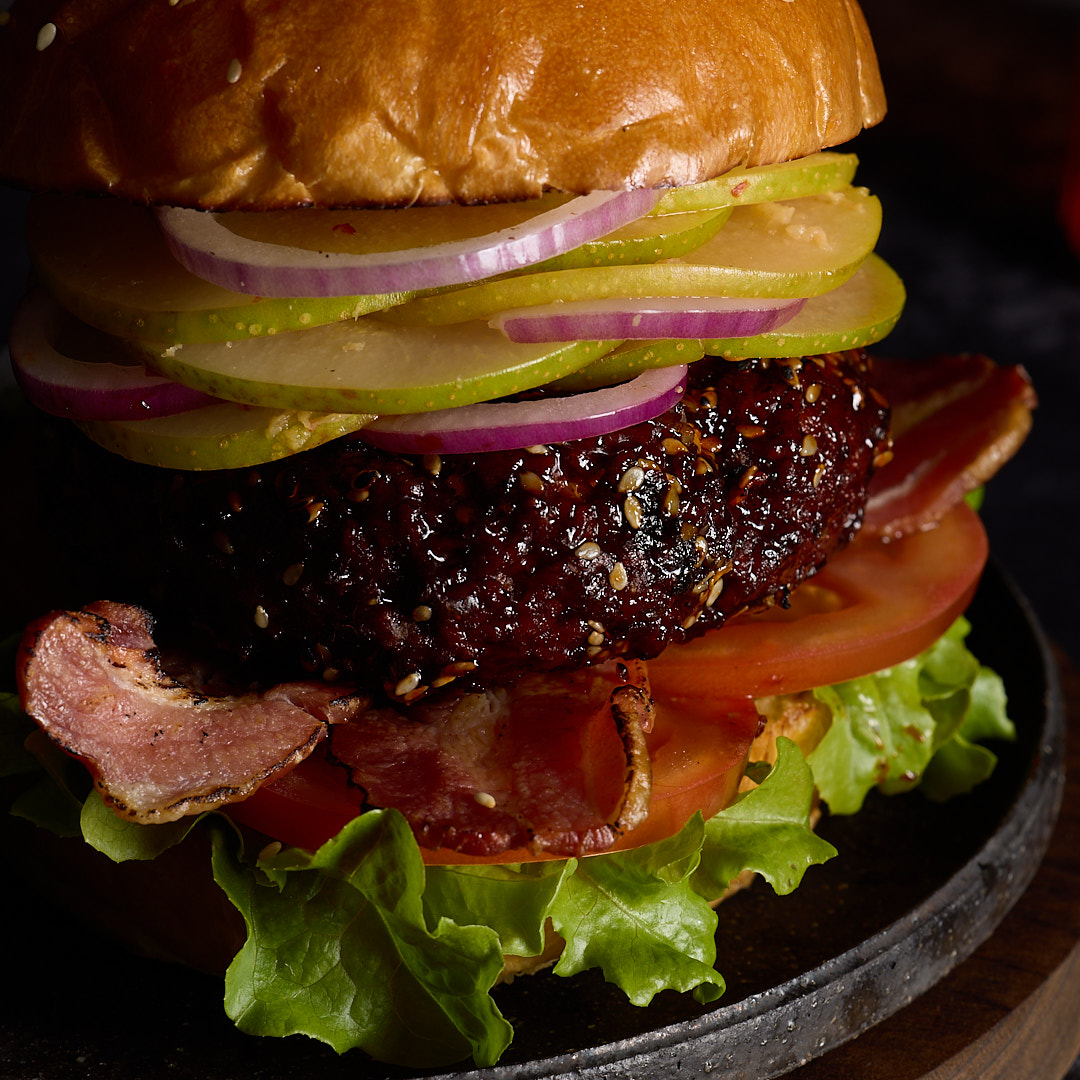 Korean-style pork burger with gochujang glazed pork patty, fresh nashi kimchi, red onion, streaky bacon, lettuce and tomato on a brioche bun with sesame seeds, stacked high on a dark wooden board against a dark moody background