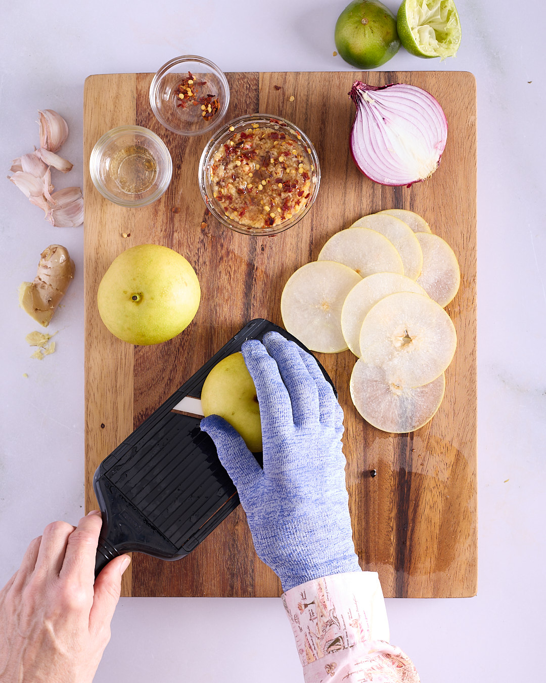 Sandra slicing nashi pear being on a handheld mandolin with a cut-resistant glove, thin rounds fanned on a wooden board alongside kimchi seasoning, red onion, garlic, ginger, lime and chilli flakes.