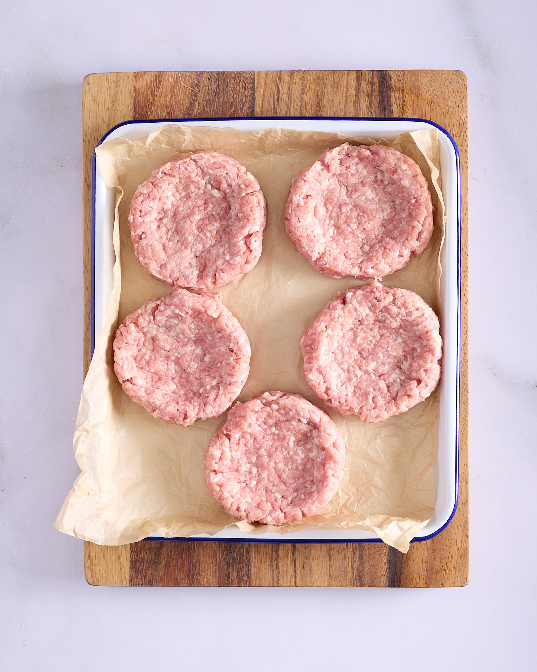 5 raw gochujang pork burger patties in a baking paper lined enamel tray on a wooden board, ready to chill before cooking.