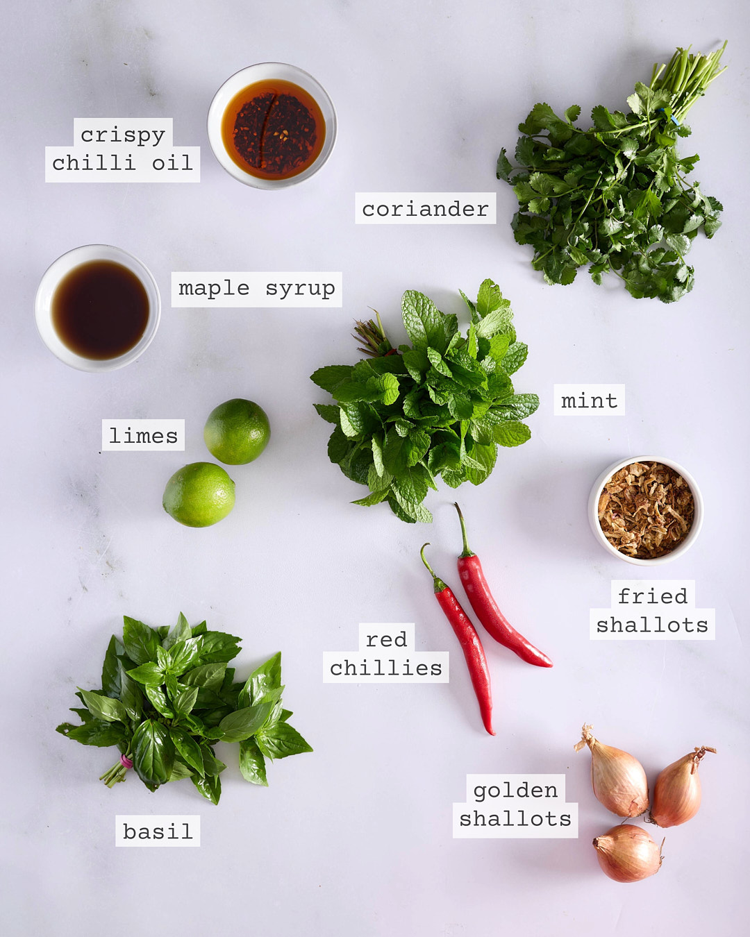 Flat lay of ingredients for the herb salad and assembly including crispy chilli oil, maple syrup, limes, mint, coriander, fried shallots, red chillies, basil and golden shallots, each clearly labelled on a white marble surface.