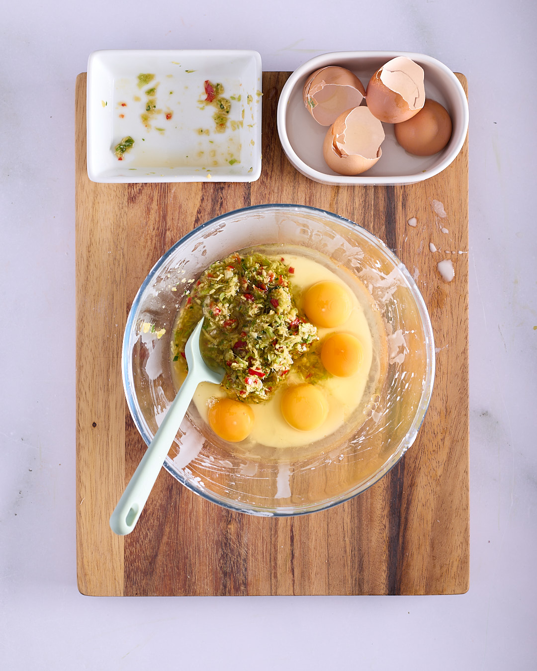 Completed Thai-style potato waffle mix in a glass bowl, grated Sebago potato combined with aromatic mixture, egg yolks and potato starch, flecked with red chilli and green herbs, with a measuring cup alongside on a wooden board.