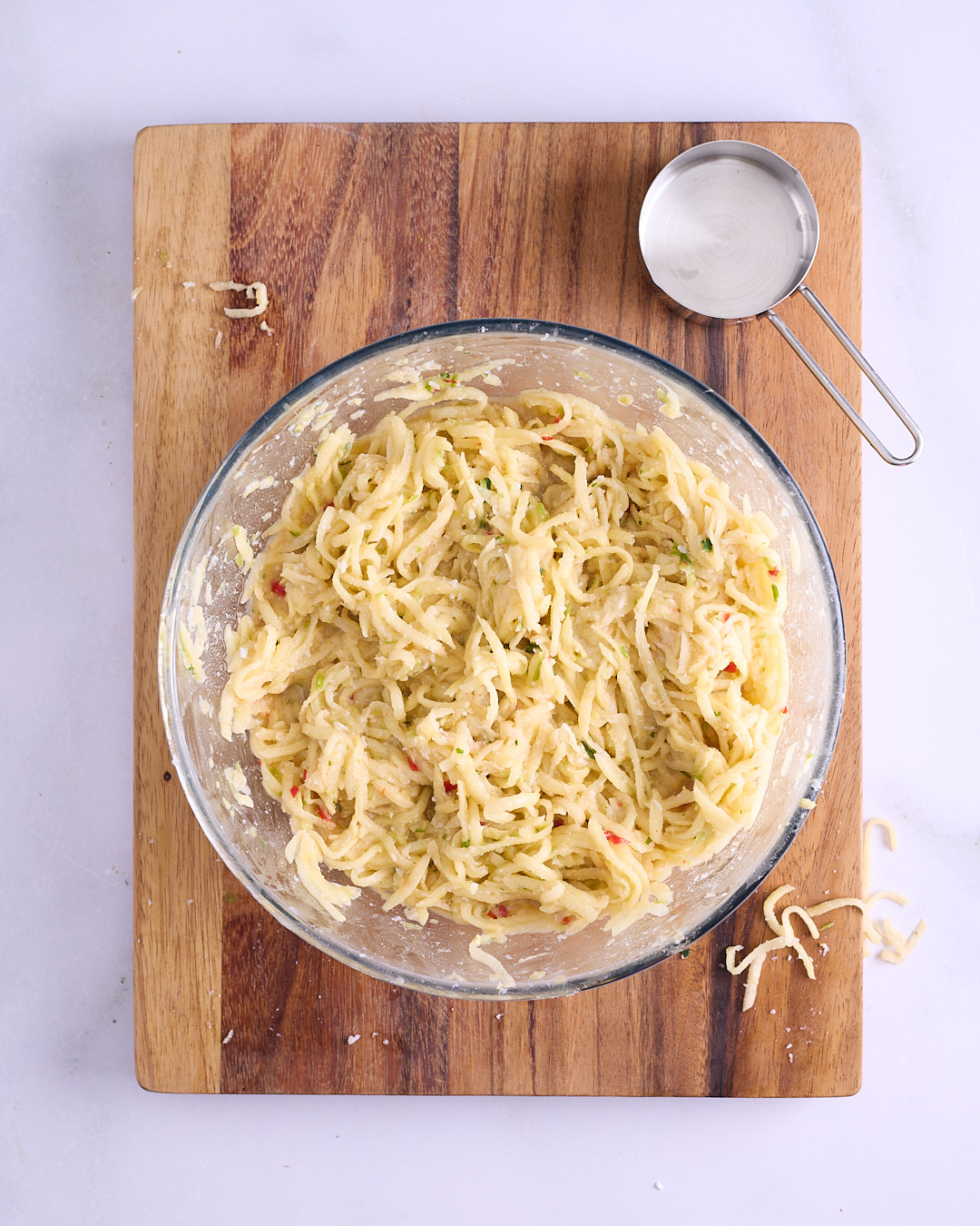 Completed Thai-style potato waffle mix in a glass bowl, grated Sebago potato combined with aromatic mixture, egg yolks and potato starch, flecked with red chilli and green herbs, with a measuring cup alongside on a wooden board.