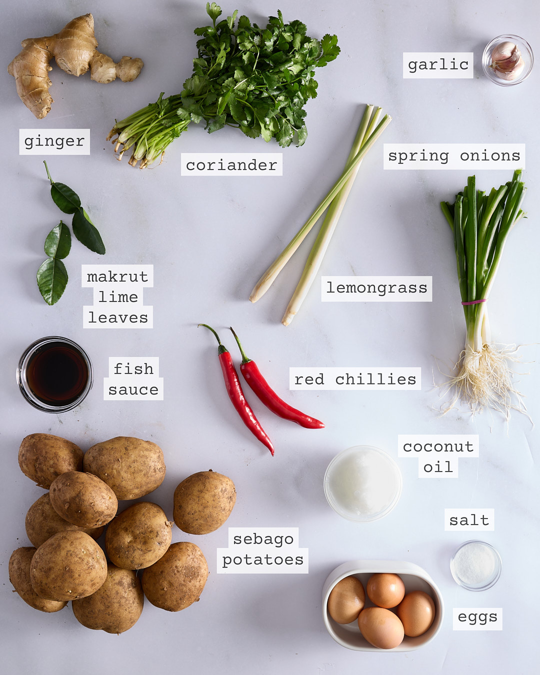 Flat lay of ingredients for Thai-style potato waffles including Sebago potatoes, ginger, coriander, spring onions, lemongrass, garlic, makrut lime leaves, red chillies, fish sauce, coconut oil, salt and eggs, each clearly labelled on a white marble surface.