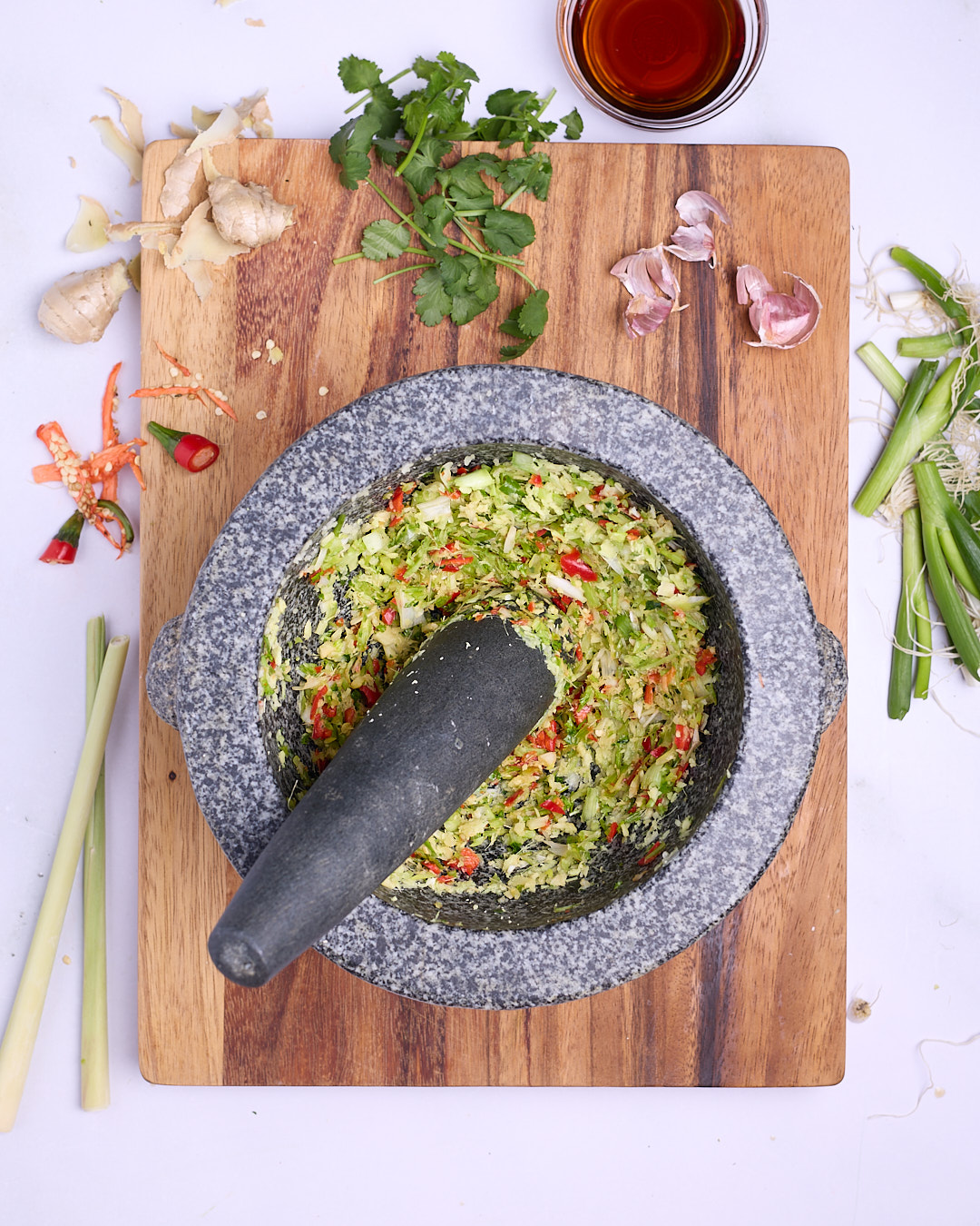 Lemongrass, ginger, garlic, chilli, coriander and spring onion roughly pounded into an aromatic mixture in a mortar and pestle on a wooden board, with lemongrass stalks and fresh coriander alongside.