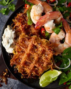 Thai-style potato waffles with fresh cooked prawns, fried shallot tartare, Thai herb salad and maple chilli oil, served on a dark plate with lime wedges, red onion and fresh coriander.