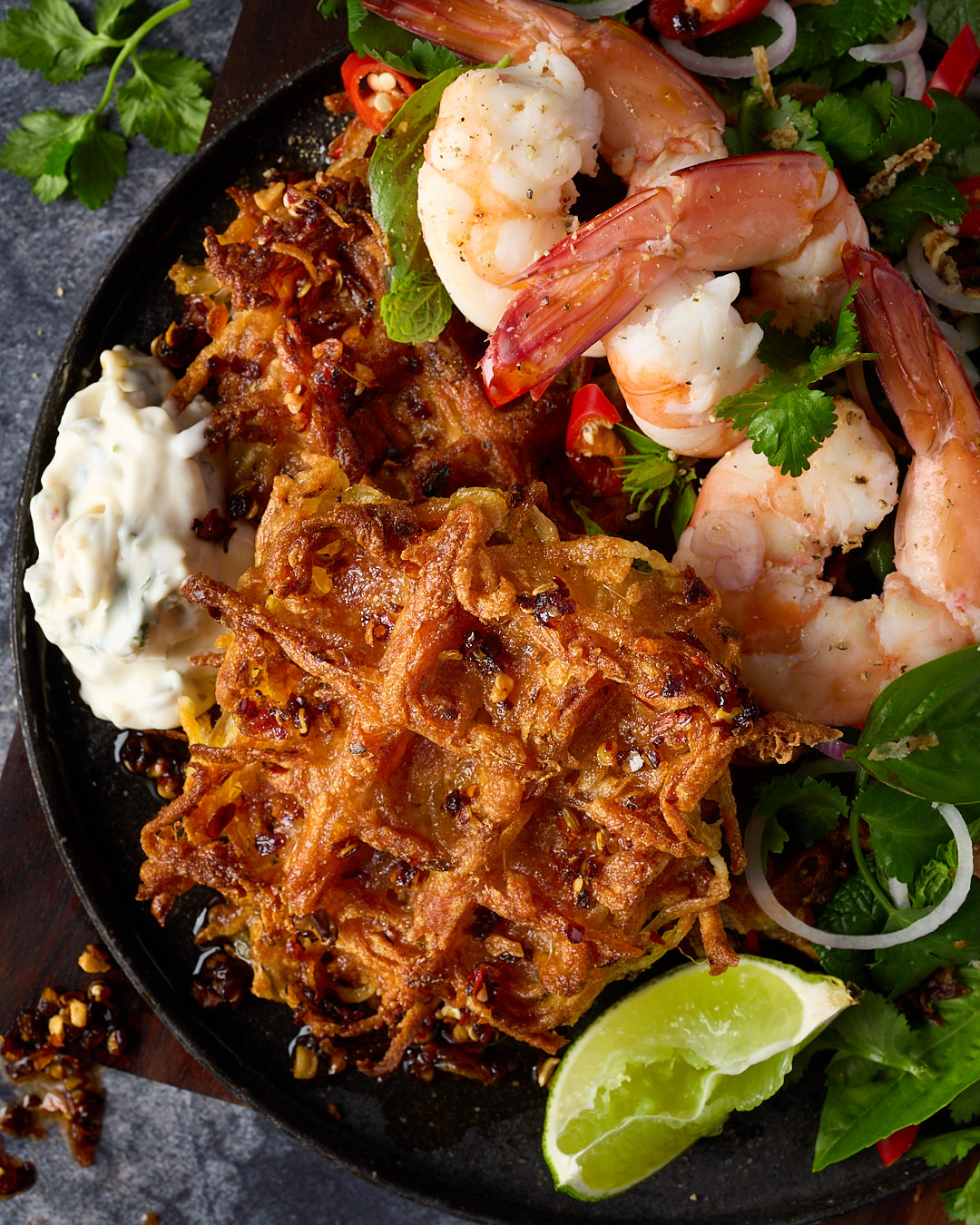 Thai-style potato waffles with fresh cooked prawns, fried shallot tartare, Thai herb salad and maple chilli oil, served on a dark plate with lime wedges, red onion and fresh coriander.