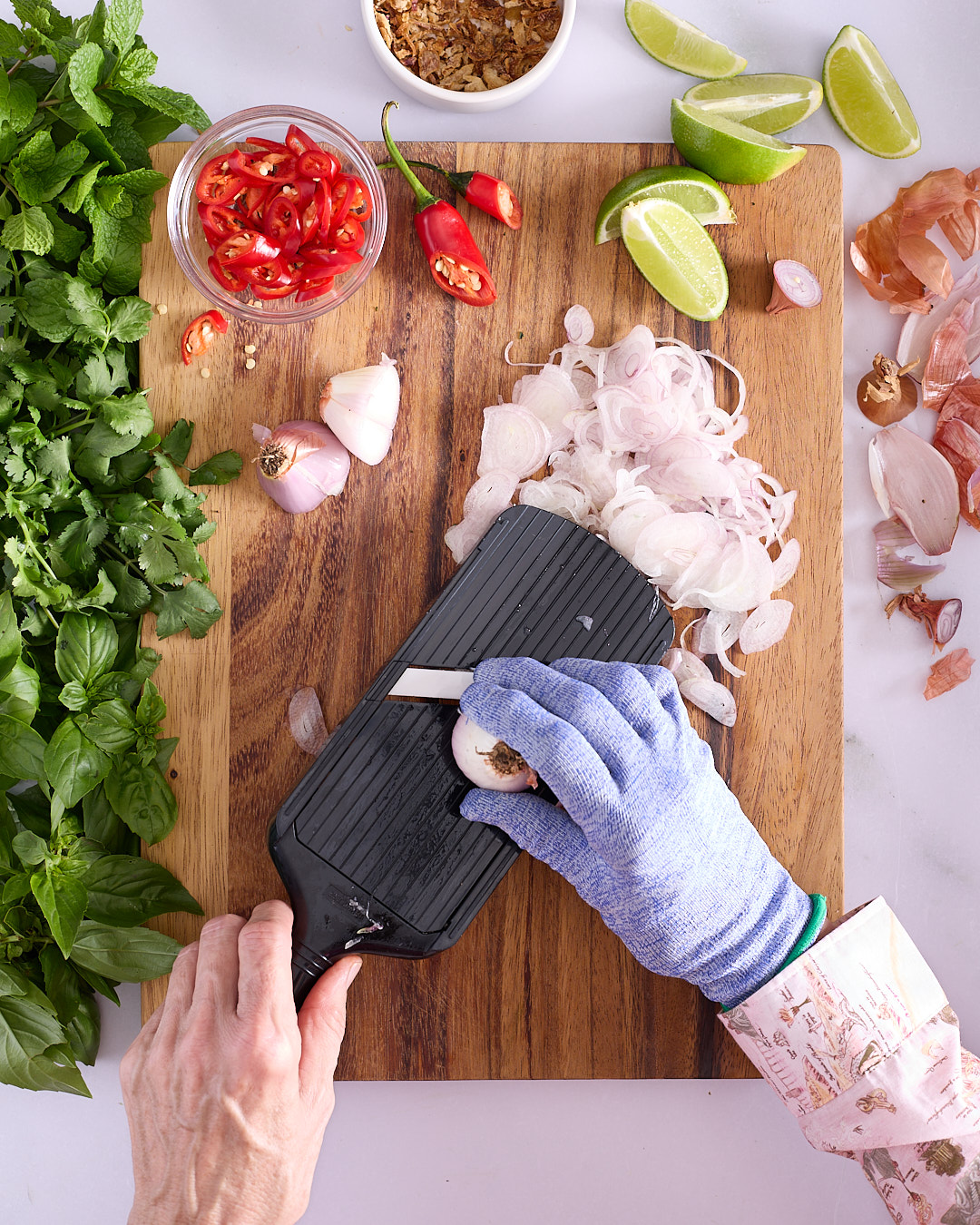 Sandra slicing golden shallots on a mandolin with a cut-resistant glove on a wooden board, with sliced red chilli, fresh mint, coriander, basil, fried shallots and lime wedges alongside.