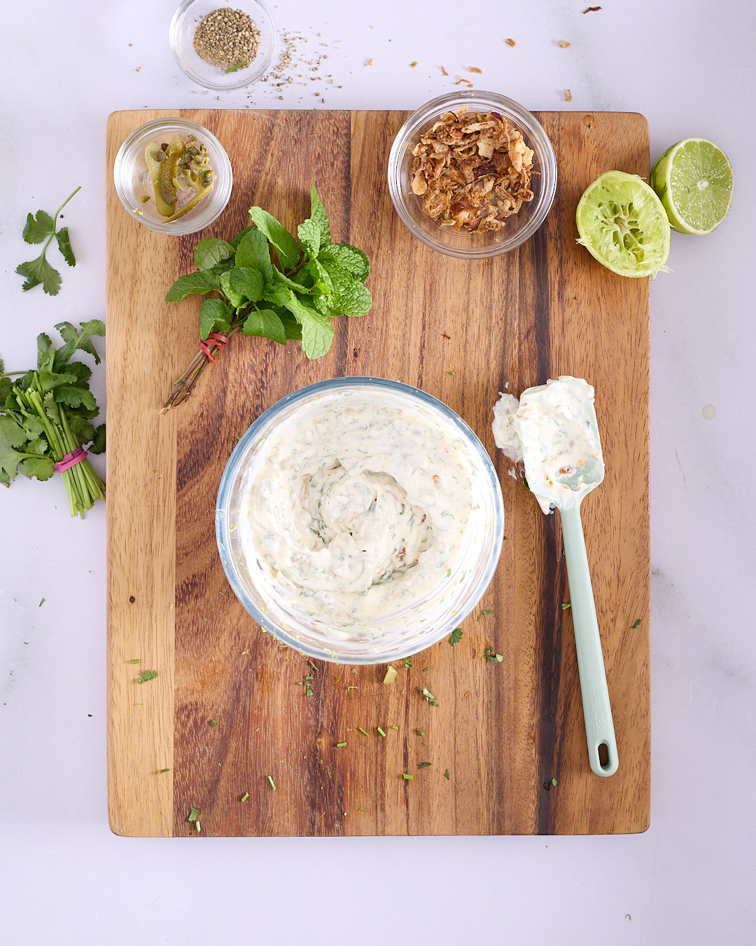 Overhead view of a wooden cutting board with a bowl of prepared fried shallot tartare. The creamy mixture is flecked with chopped herbs and pieces of cornichons and fried shallots. Surrounding the bowl are fresh mint and coriander, halved limes, small bowls of white pepper, sliced cornichons, and crispy fried shallots, with a spatula coated in the finished tartare resting beside the bowl.
