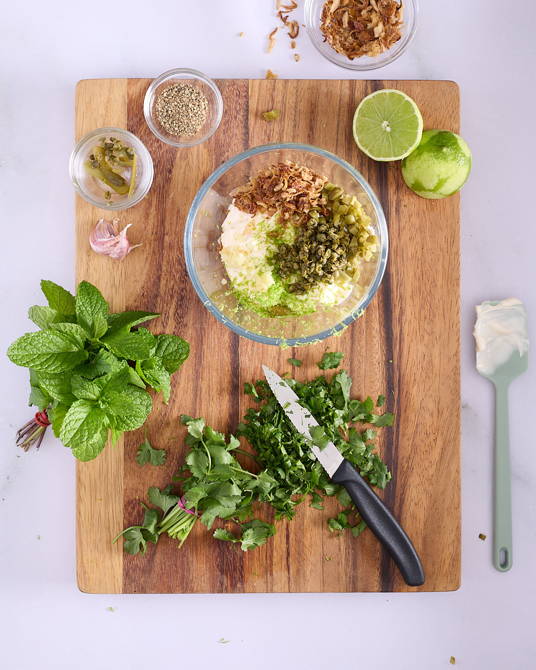 Overhead view of a wooden cutting board with ingredients for a mayonnaise tartare. A glass bowl contains mayonnaise topped with chopped capers, diced cornichons, crispy fried shallots, minced garlic, and lime zest. Around the bowl are fresh mint and coriander, a halved lime, garlic cloves, chopped herbs with a knife, small bowls of sesame seeds, sliced pickles, and fried onions, plus a spatula with a dollop of mayonnaise.