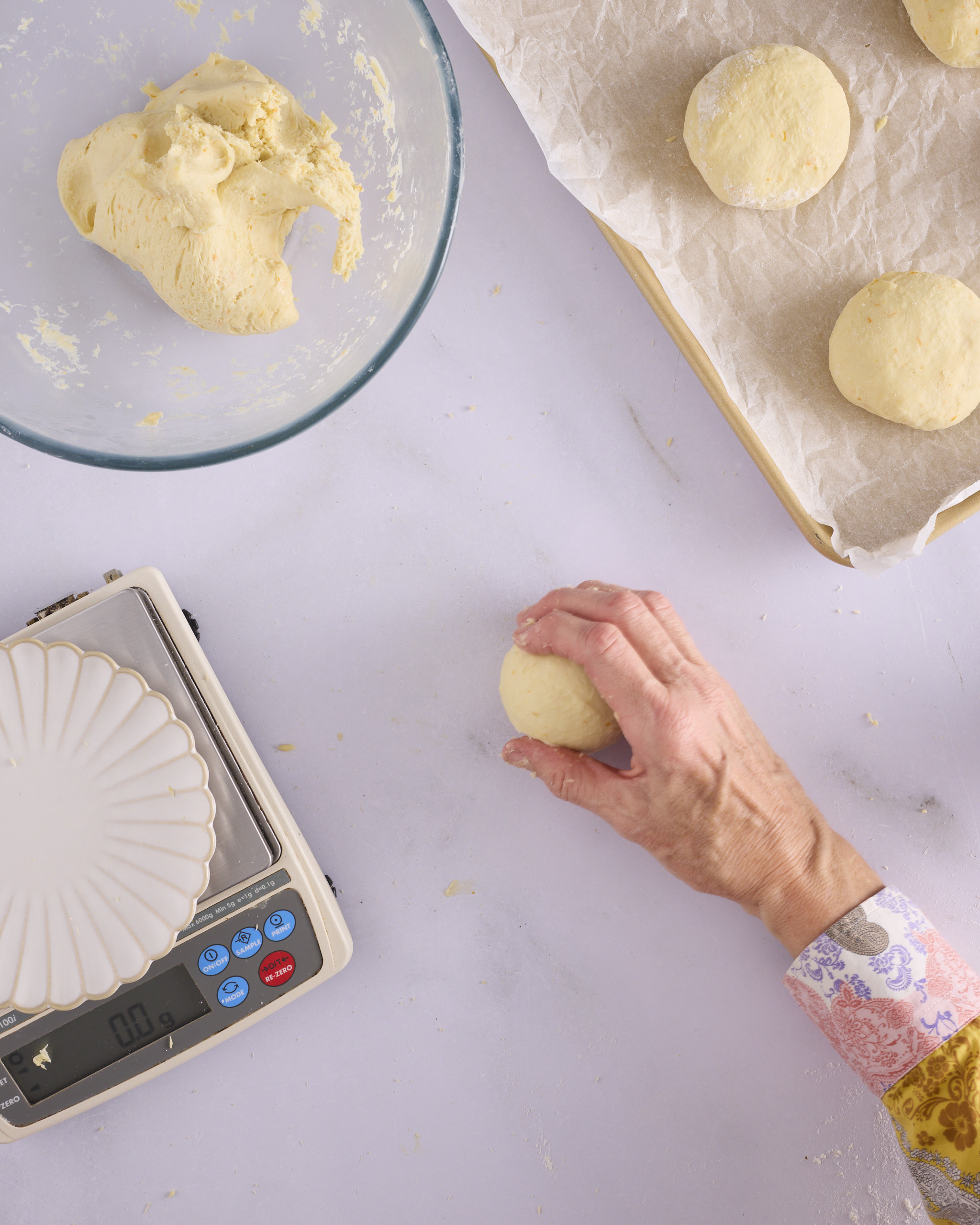 Sandra rolls a 100g dough ball for blueberry cheesecake kolache in small circles on the bench to tighten the surface, with digital scales and a lined tray of shaped balls alongside.