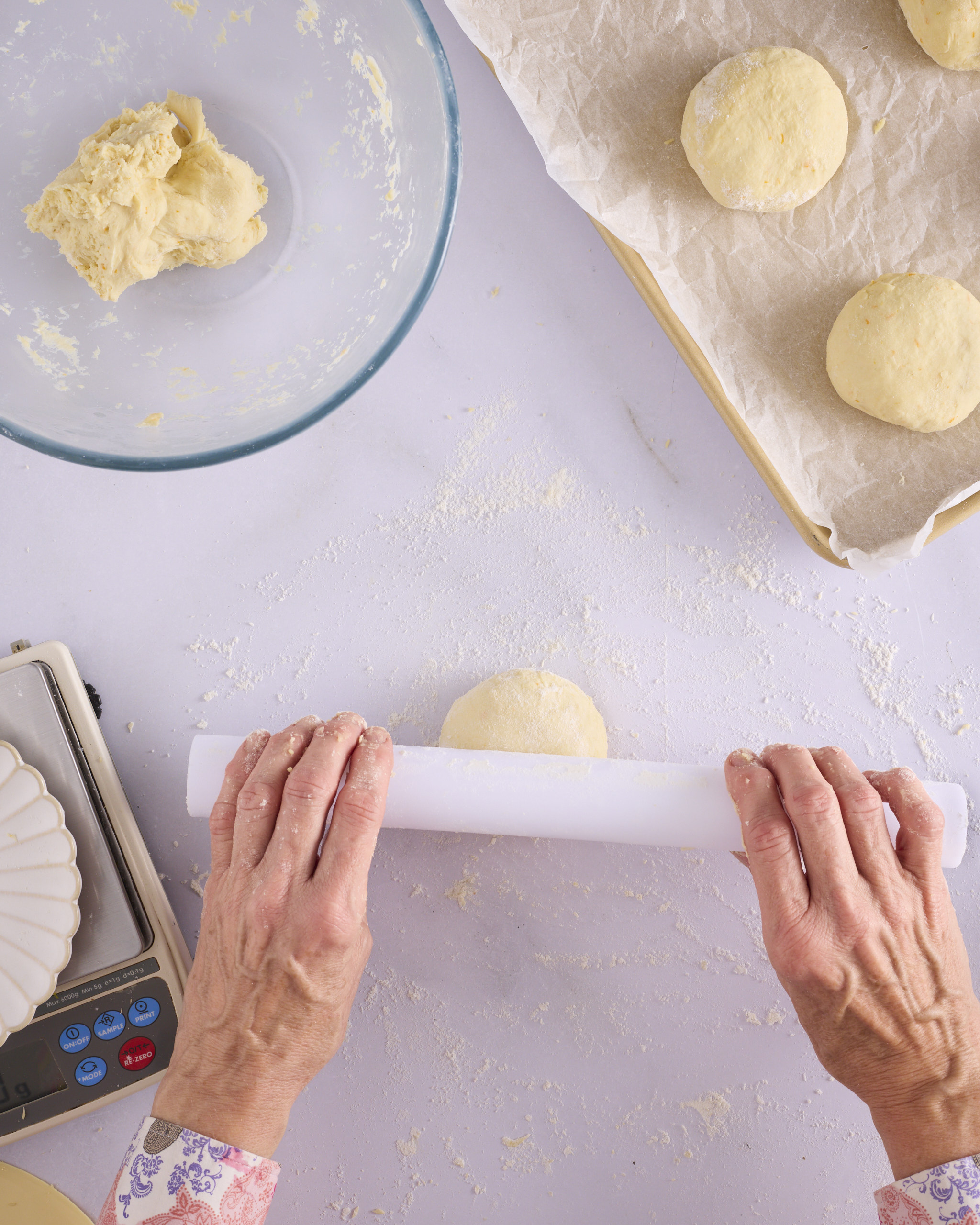 Sandra uses a small rolling pin flattens a shaped dough ball for blueberry cheesecake kolache into a disc on a lightly floured bench, with the bowl of dough and a lined tray alongside.