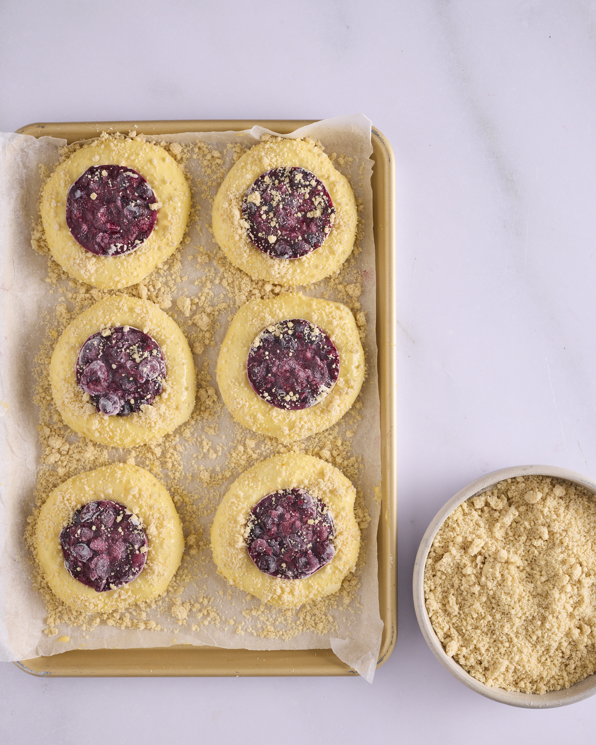 Six blueberry cheesecake kolache assembled and ready to bake on a lined gold baking tray. A frozen insert of blueberry rosemary jam over cheesecake is pressed into the centre of each bun, with cinnamon crumble scattered over the exposed dough and across the paper. A bowl of cinnamon crumble sits beside the tray.