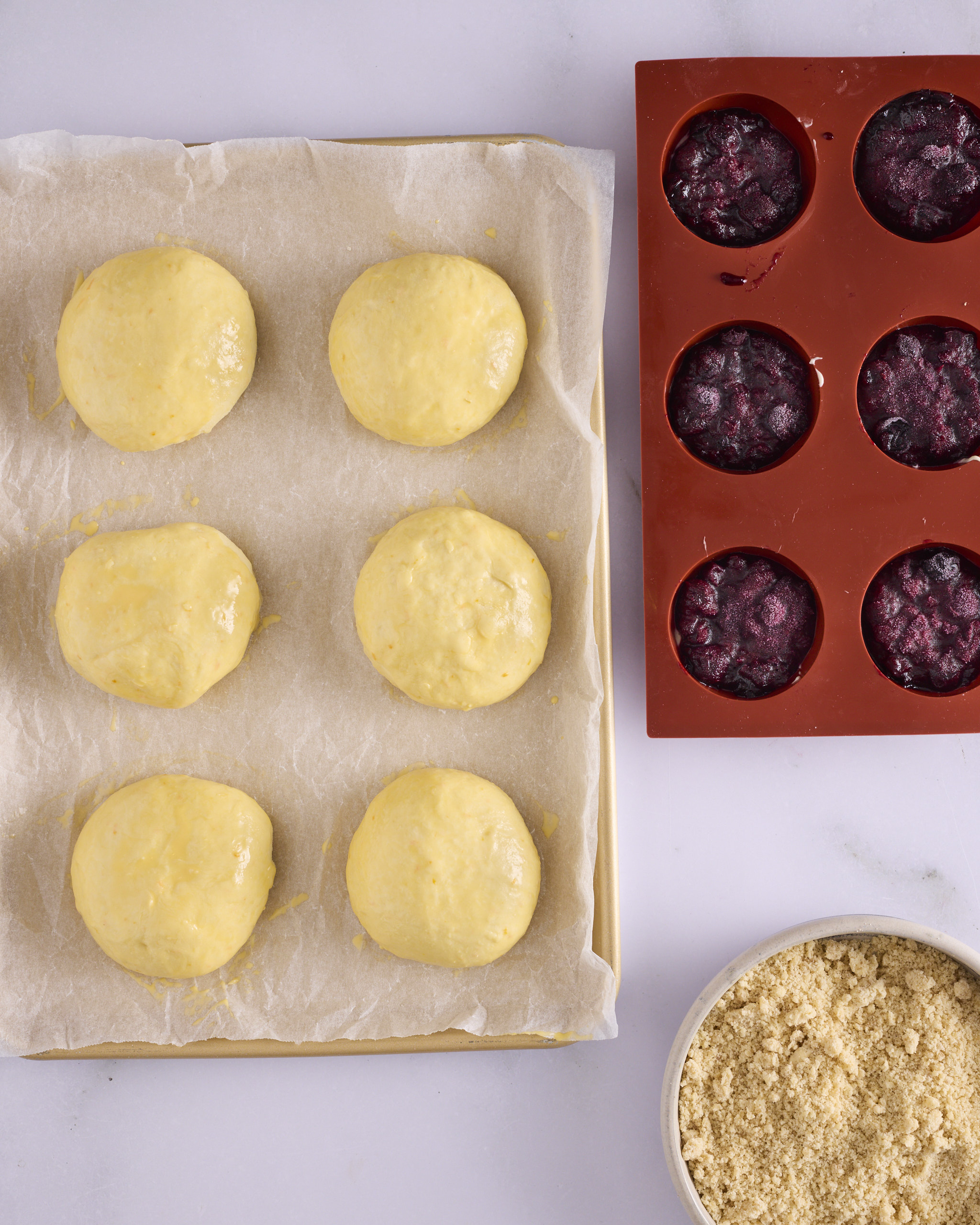 Six proved dough discs for blueberry cheesecake kolache on a lined gold baking tray, brushed with egg wash and risen to just under double. A silicone mould of frozen blueberry rosemary jam and cheesecake inserts sits alongside with a bowl of cinnamon crumble.