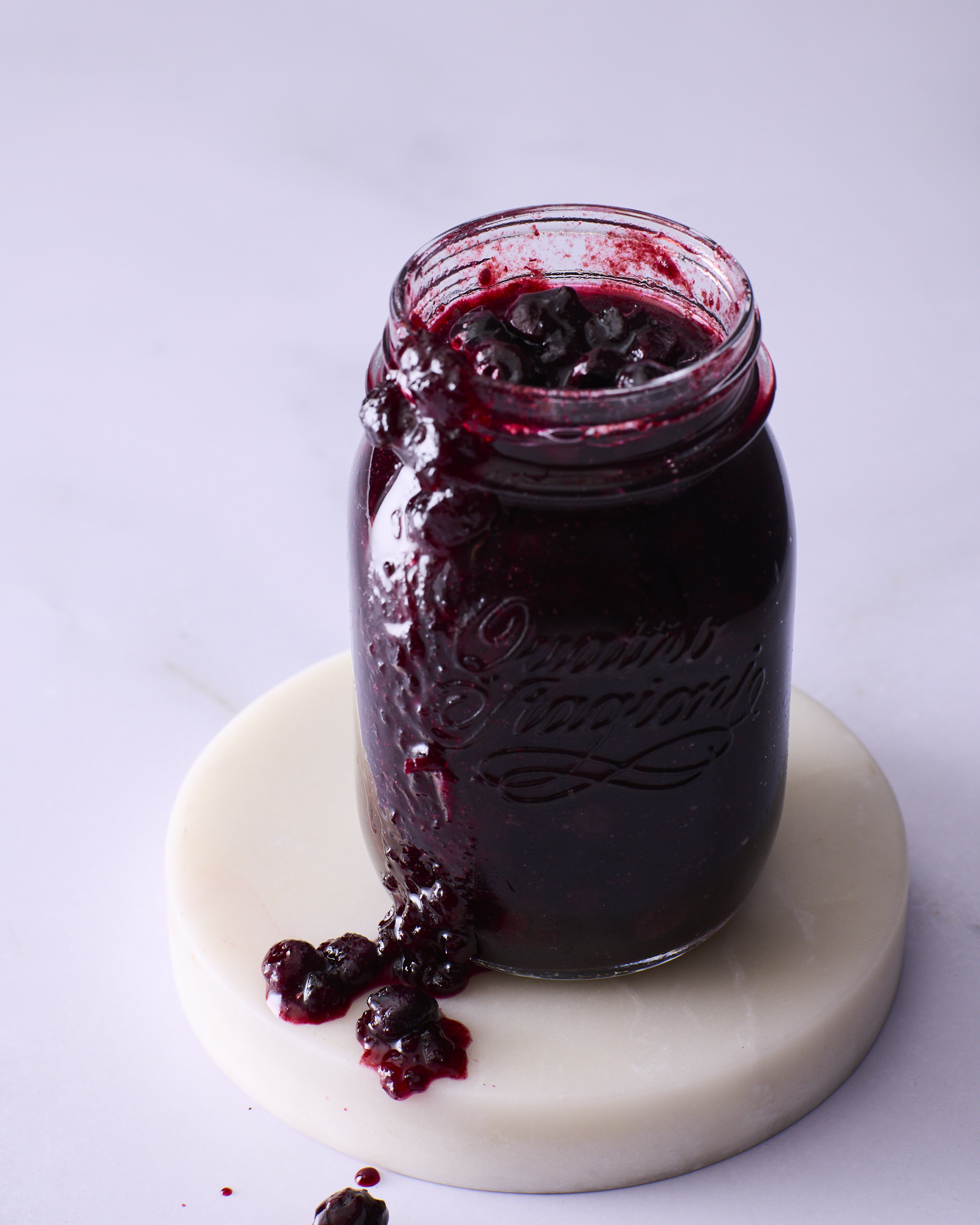 An open jar of blueberry rosemary jam for blueberry cheesecake kolache, standing on a white marble coaster. The jam is dark and glossy with whole blueberries visible at the rim, and it has run down the side of the jar onto the marble.