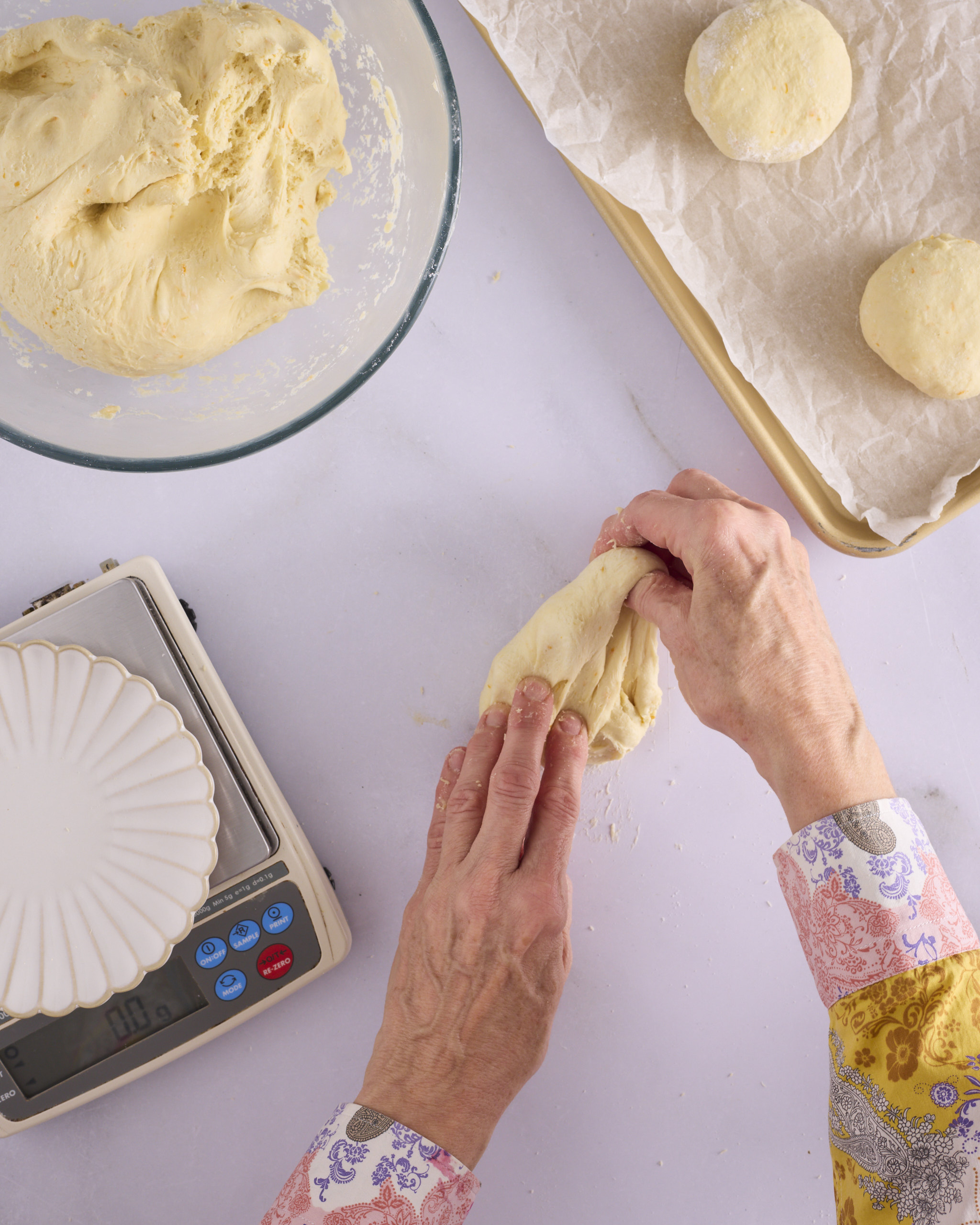 Sandra stretches a corner of a 100g piece of foundation sweet dough for blueberry cheesecake kolache on the bench, with digital scales alongside and two shaped dough balls on a lined baking tray.