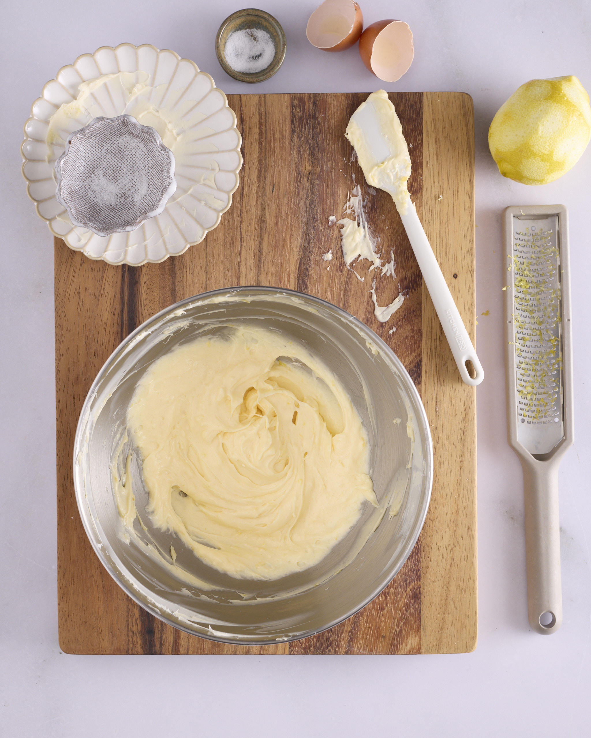 The finished lemon cheesecake filling for blueberry cheesecake kolache in a stand mixer bowl, smooth and pale yellow with flecks of lemon zest through it. A spatula, a zested lemon and a Microplane sit on the wooden board beside the bowl.