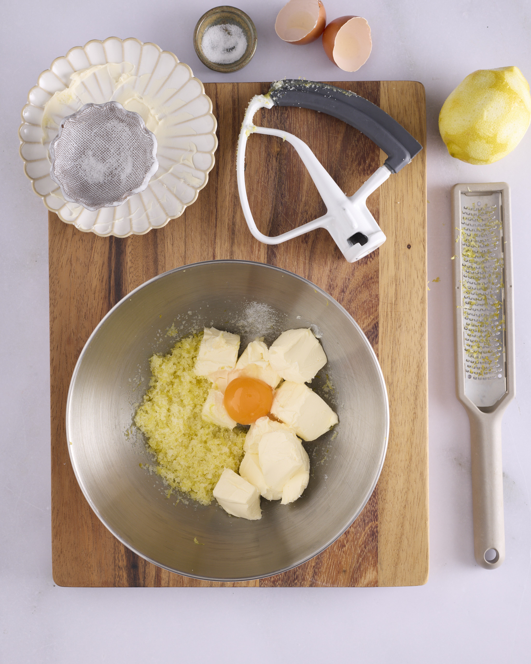 Ingredients for the lemon cheesecake filling for blueberry cheesecake kolache in a stand mixer bowl on a wooden board. Lemon zest rubbed into white sugar sits alongside cubes of Philadelphia cream cheese and an egg yolk, with salt, a zested lemon, a Microplane and the paddle attachment beside the bowl.
