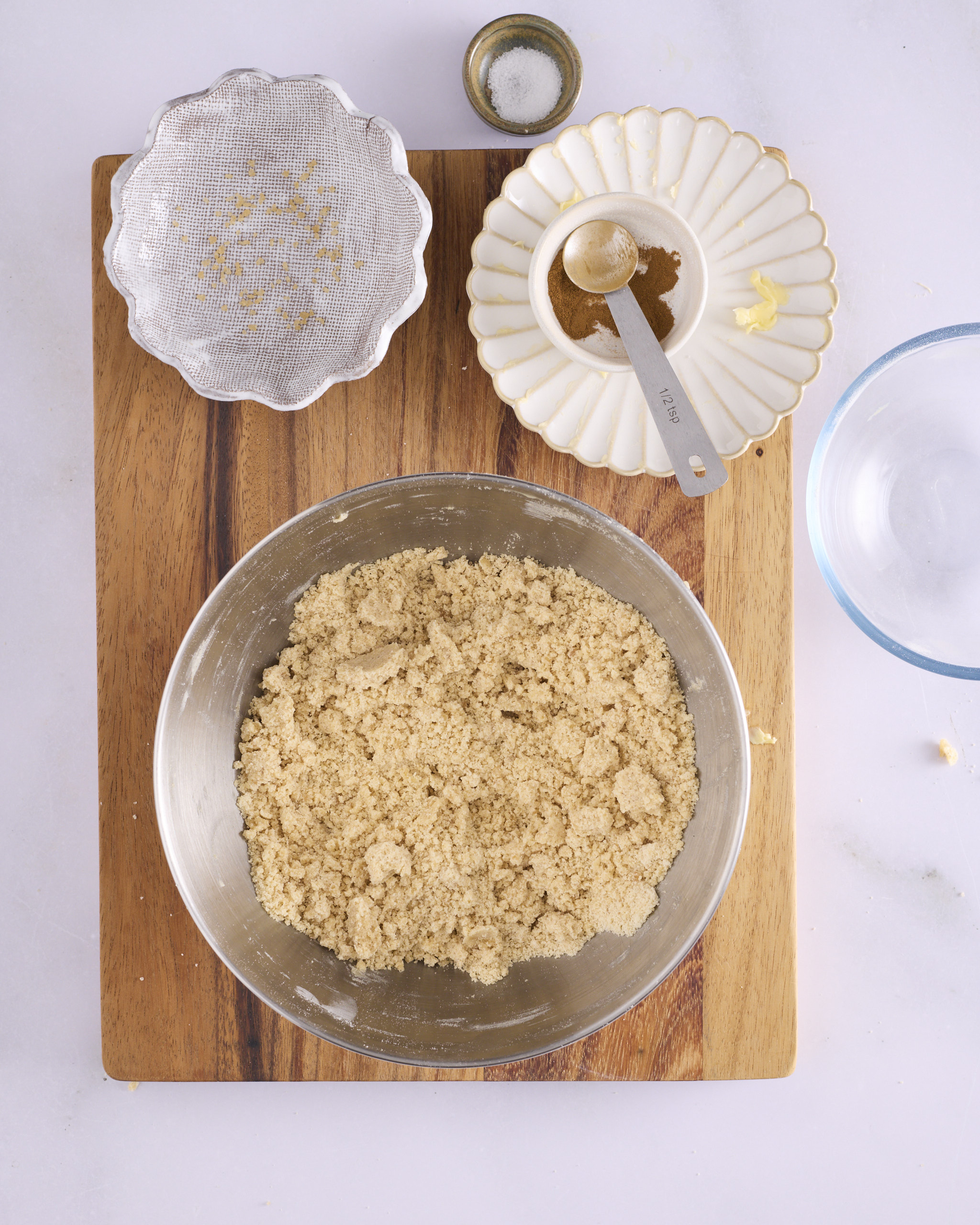 Cinnamon crumble for blueberry cheesecake kolache in a stand mixer bowl on a wooden board, rubbed to a coarse sandy texture. A sifter, a small bowl of salt and a dish of cinnamon with a half teaspoon measure sit beside it.