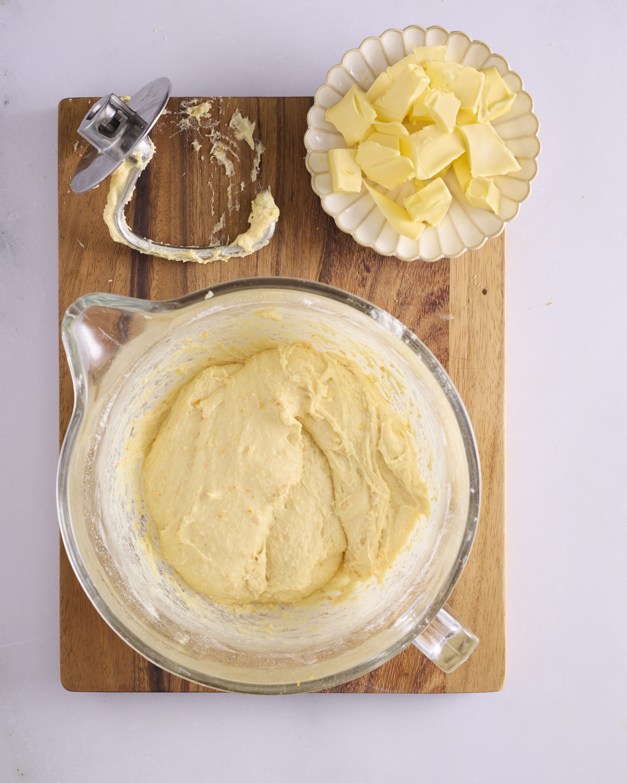 Foundation sweet dough in a glass stand mixer bowl on a wooden board, soft and slack with flecks of orange zest through it. The dough hook rests on the board beside a dish of cubed unsalted butter waiting to be added.