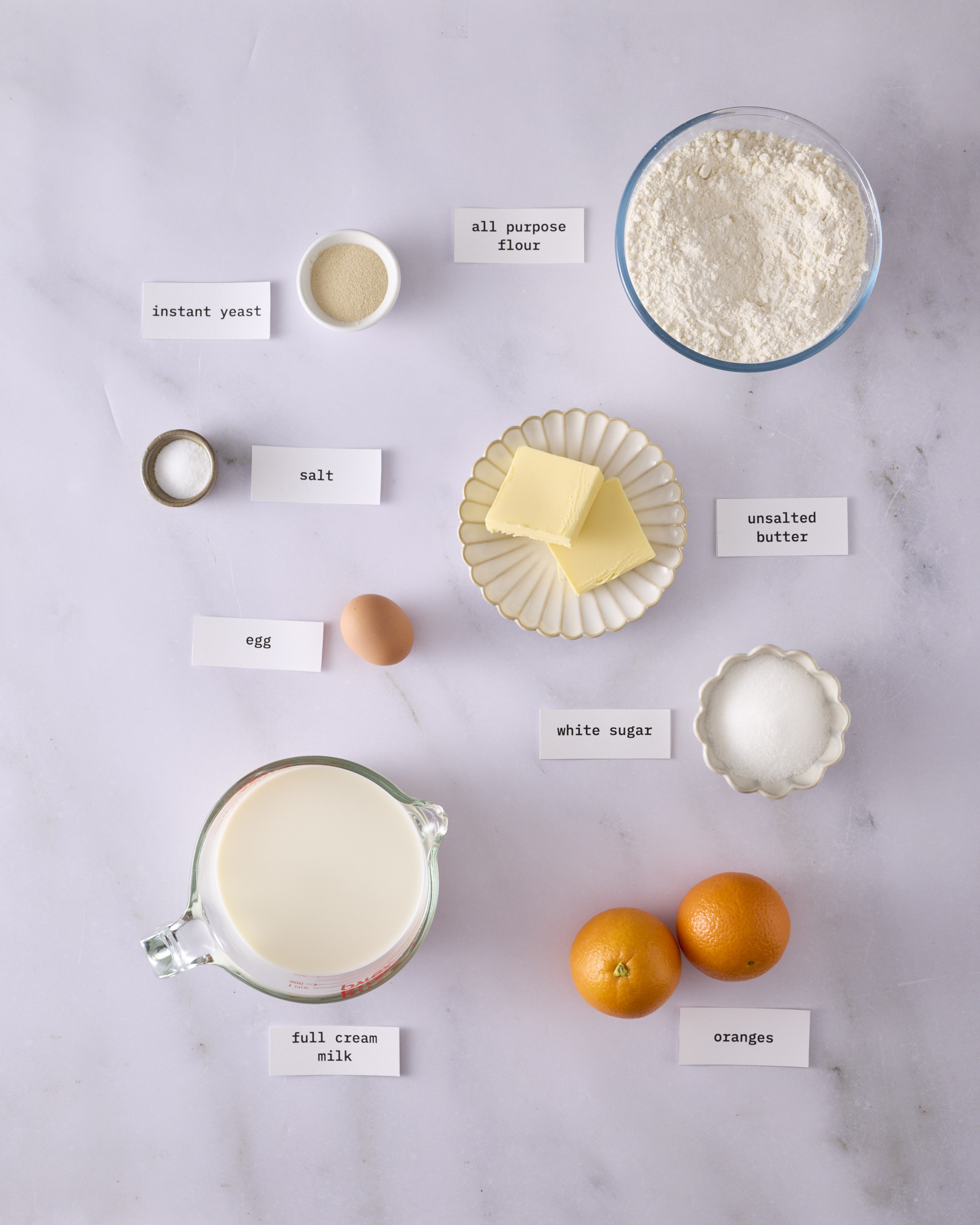 Labelled ingredients for the foundation sweet dough laid out on a marble surface. All-purpose flour, full cream milk, unsalted butter, instant yeast, white sugar, salt, one egg and two oranges.