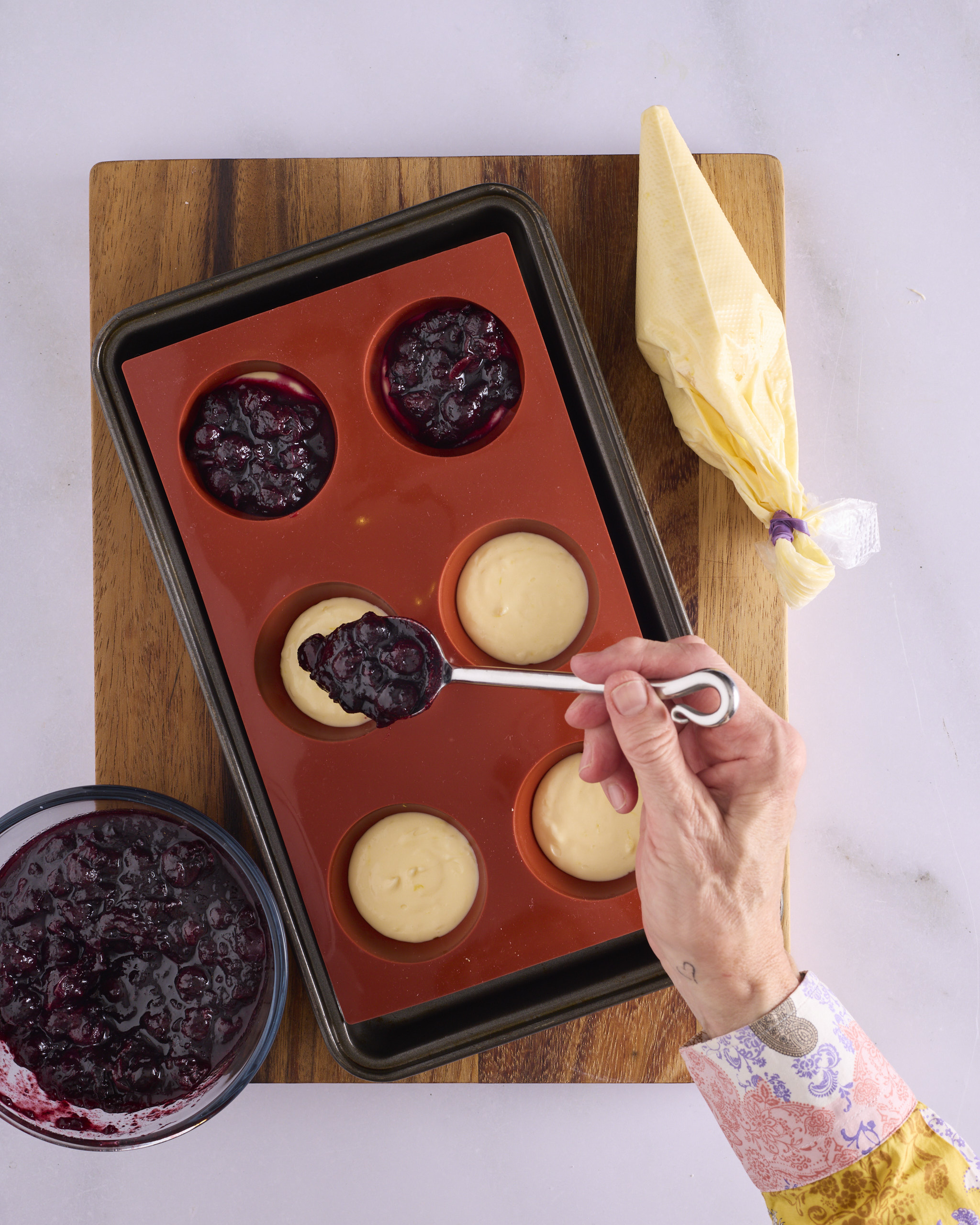 Sandra spoons blueberry rosemary jam over the lemon cheesecake layer in a silicone hemisphere mould to make the frozen filling inserts for blueberry cheesecake kolache. Two moulds are already topped with jam, three hold cheesecake waiting for it, and a piping bag of cheesecake filling and a bowl of jam sit alongside on the tray.