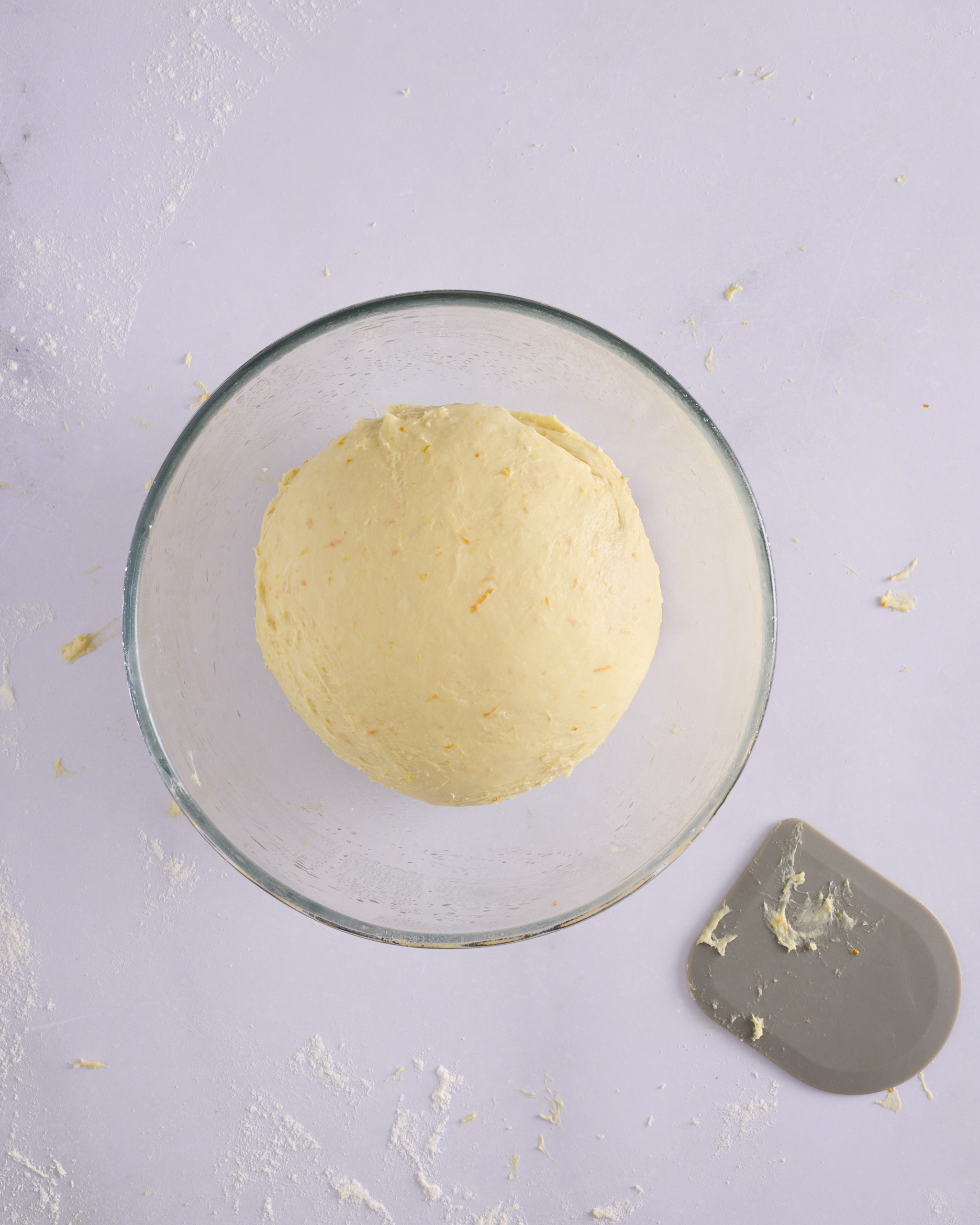 The finished foundation sweet dough resting in a greased glass bowl, smooth and taut with a rounded top. A grey dough scraper sits on the bench beside it.