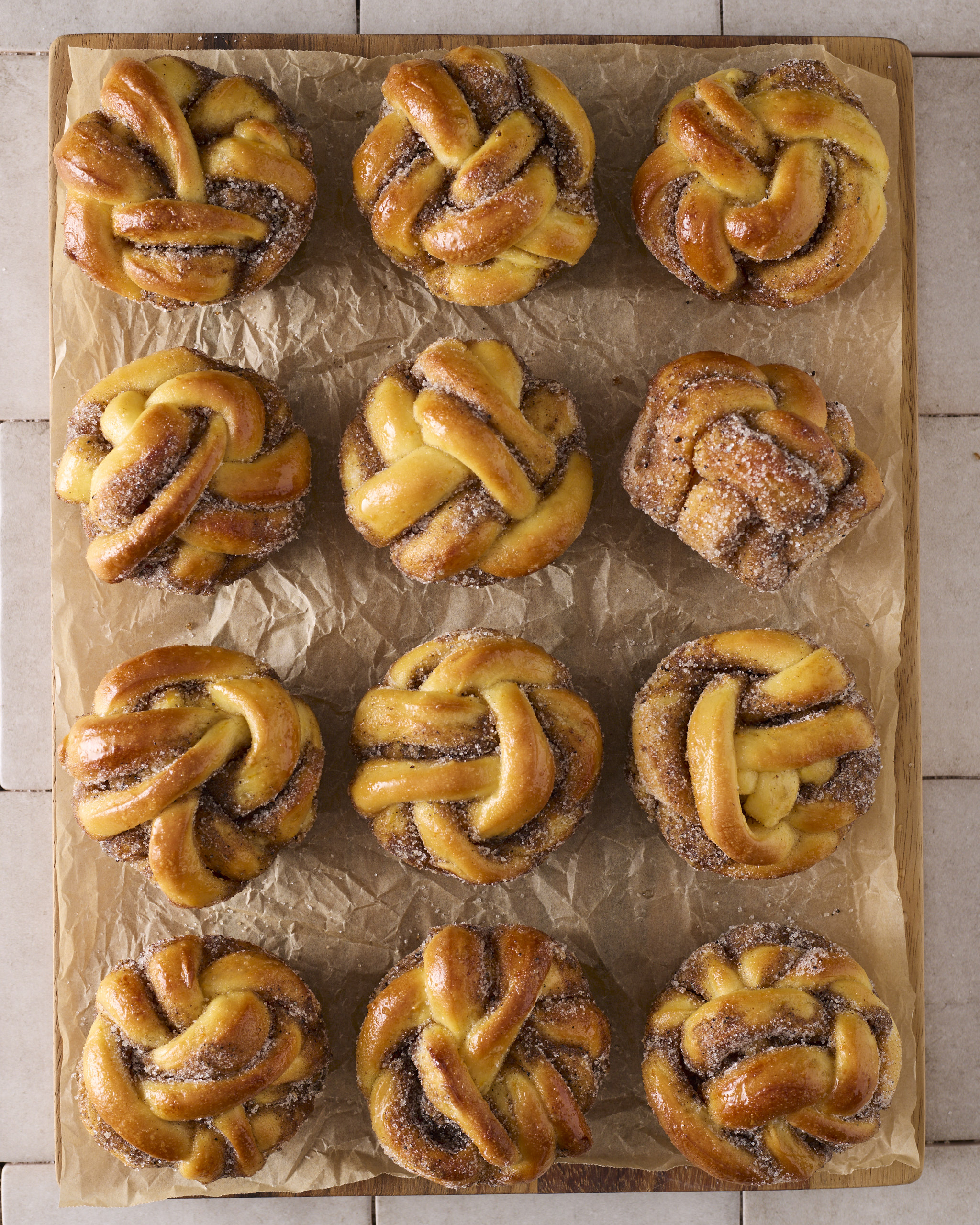 A dozen Cardamom Orange Knots arranged on parchment paper, each one twisted into a knot shape and finished with a golden, sugared crust.
