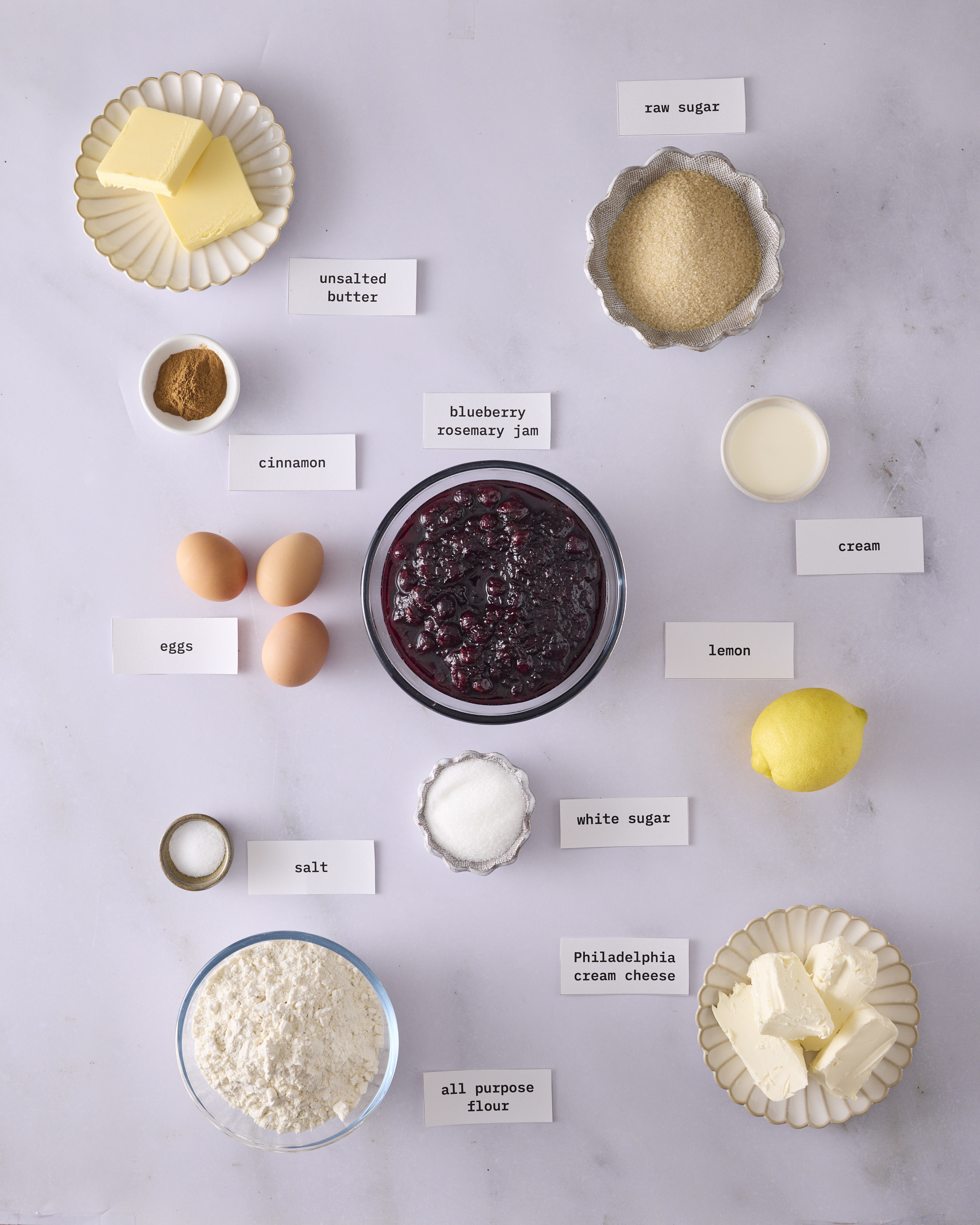 Labelled ingredients for the blueberry cheesecake kolache filling laid out on a marble surface. A bowl of blueberry rosemary jam sits in the centre with Philadelphia cream cheese, three eggs, a lemon, cream, all-purpose flour, raw sugar, white sugar, unsalted butter, cinnamon and salt for the cheesecake filling and cinnamon crumble.