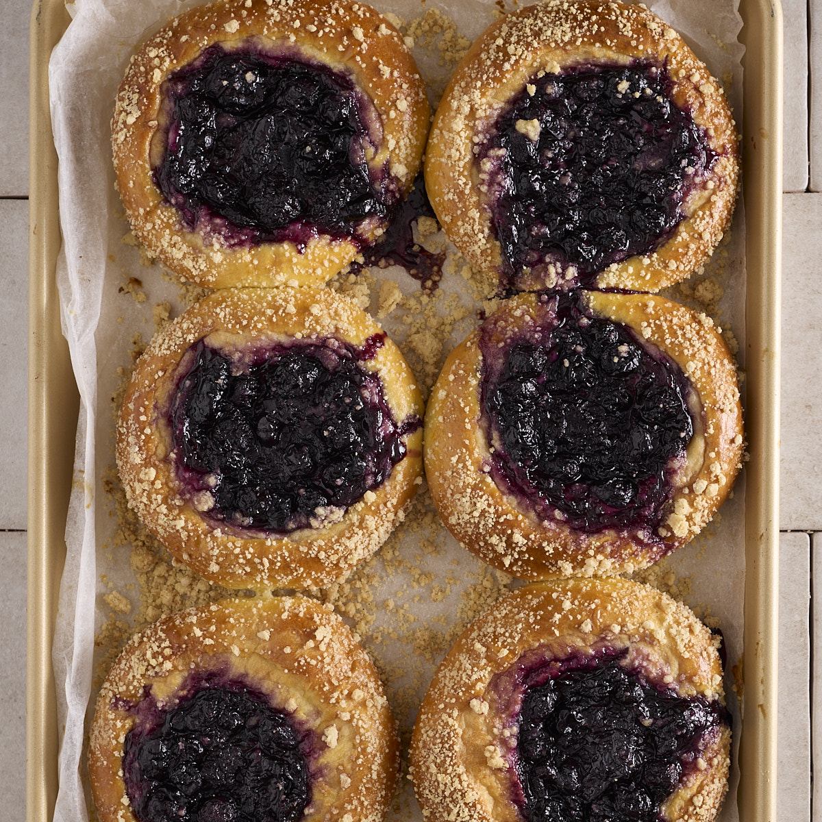 Six baked blueberry cheesecake kolache on a gold baking tray lined with baking paper. Each filled sweet bun has a centre of blueberry rosemary jam over the cheesecake filling, with cinnamon crumble scattered across the golden edges. Jam has run down the side of two of the buns onto the paper.