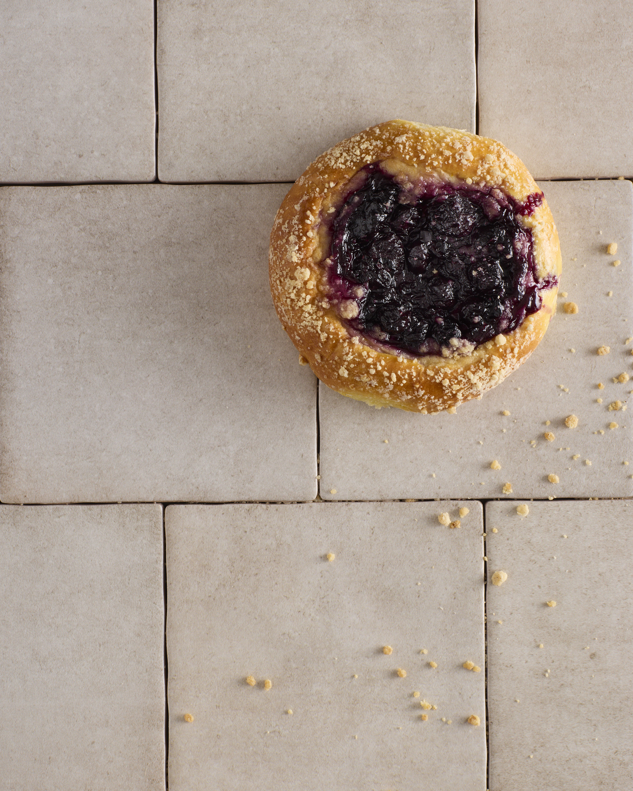 A single blueberry cheesecake kolache on a pale tiled surface, the blueberry rosemary jam glossy and dark in the centre over the cheesecake filling, with cinnamon crumble catching on the golden edge and scattered crumbs across the tiles.