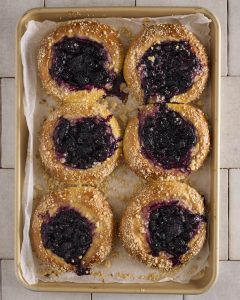 Six baked blueberry cheesecake kolache on a gold baking tray lined with baking paper. Each filled sweet bun has a centre of blueberry rosemary jam over the cheesecake filling, with cinnamon crumble scattered across the golden edges. Jam has run down the side of two of the buns onto the paper.