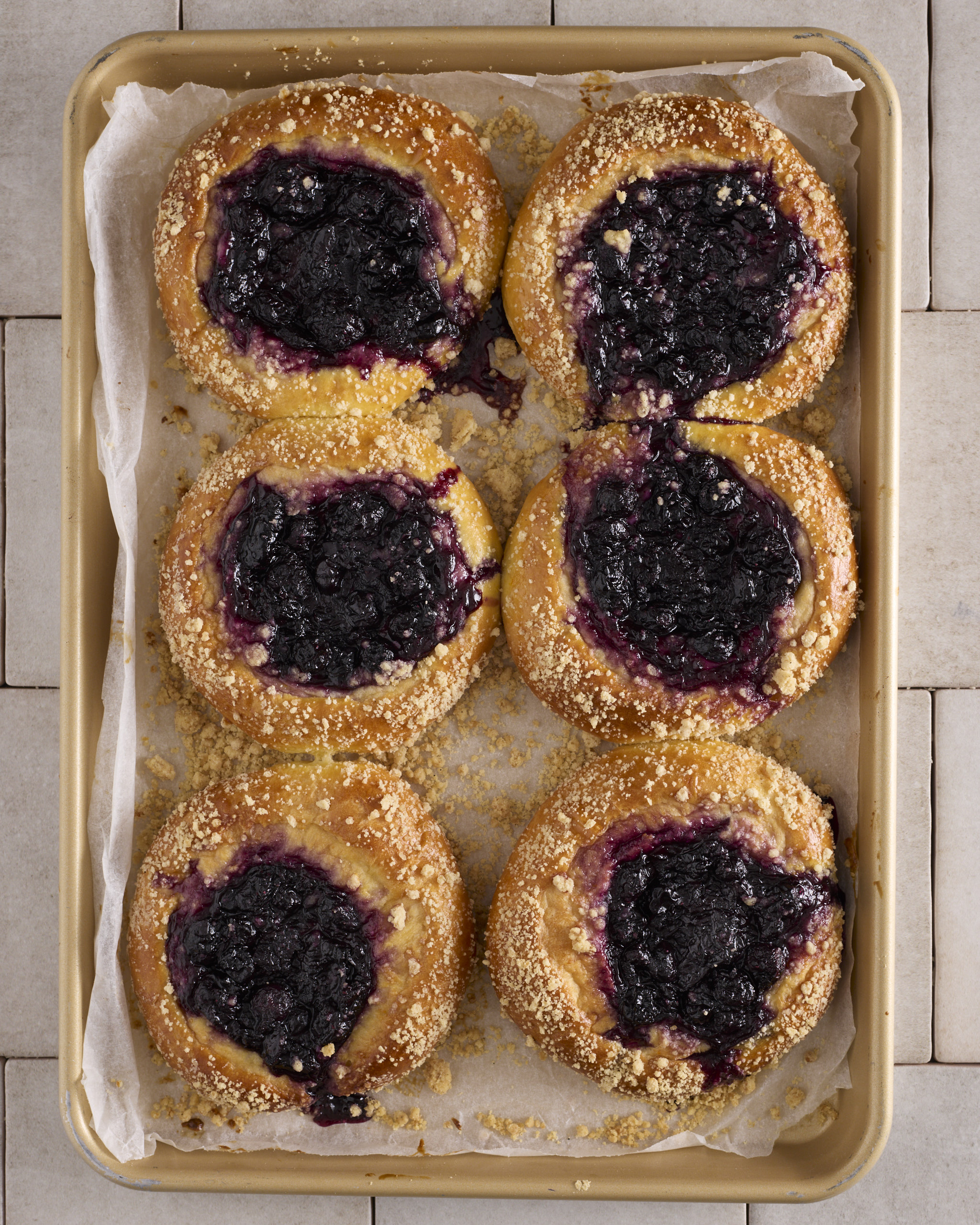 Six baked blueberry cheesecake kolache on a gold baking tray lined with baking paper. Each filled sweet bun has a centre of blueberry rosemary jam over the cheesecake filling, with cinnamon crumble scattered across the golden edges. Jam has run down the side of two of the buns onto the paper.