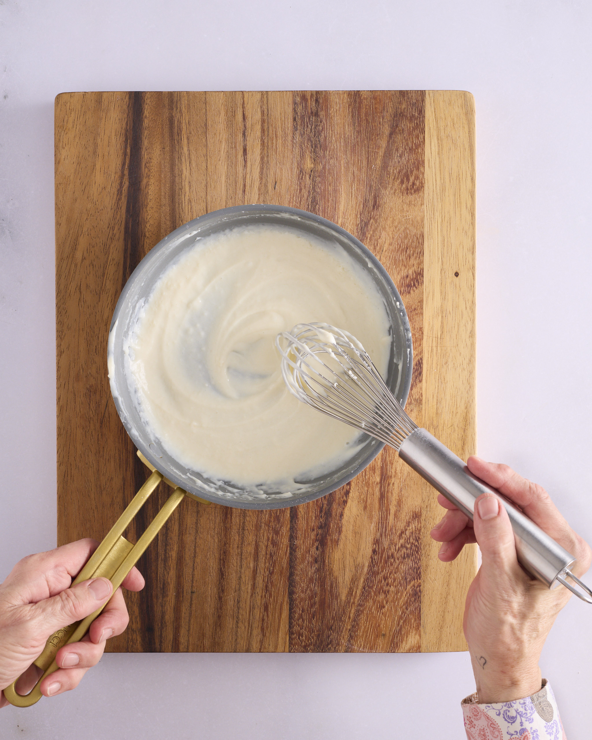 Sandra hand whisks the tangzhong paste for a foundation sweet dough recipe in a small pan set on a wooden board. The mixture is thick, pale and glossy, with the whisk leaving swirls through it.