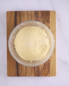 The foundation sweet dough risen in a glass bowl on a wooden board, smooth and domed, with flecks of orange zest through the surface.
