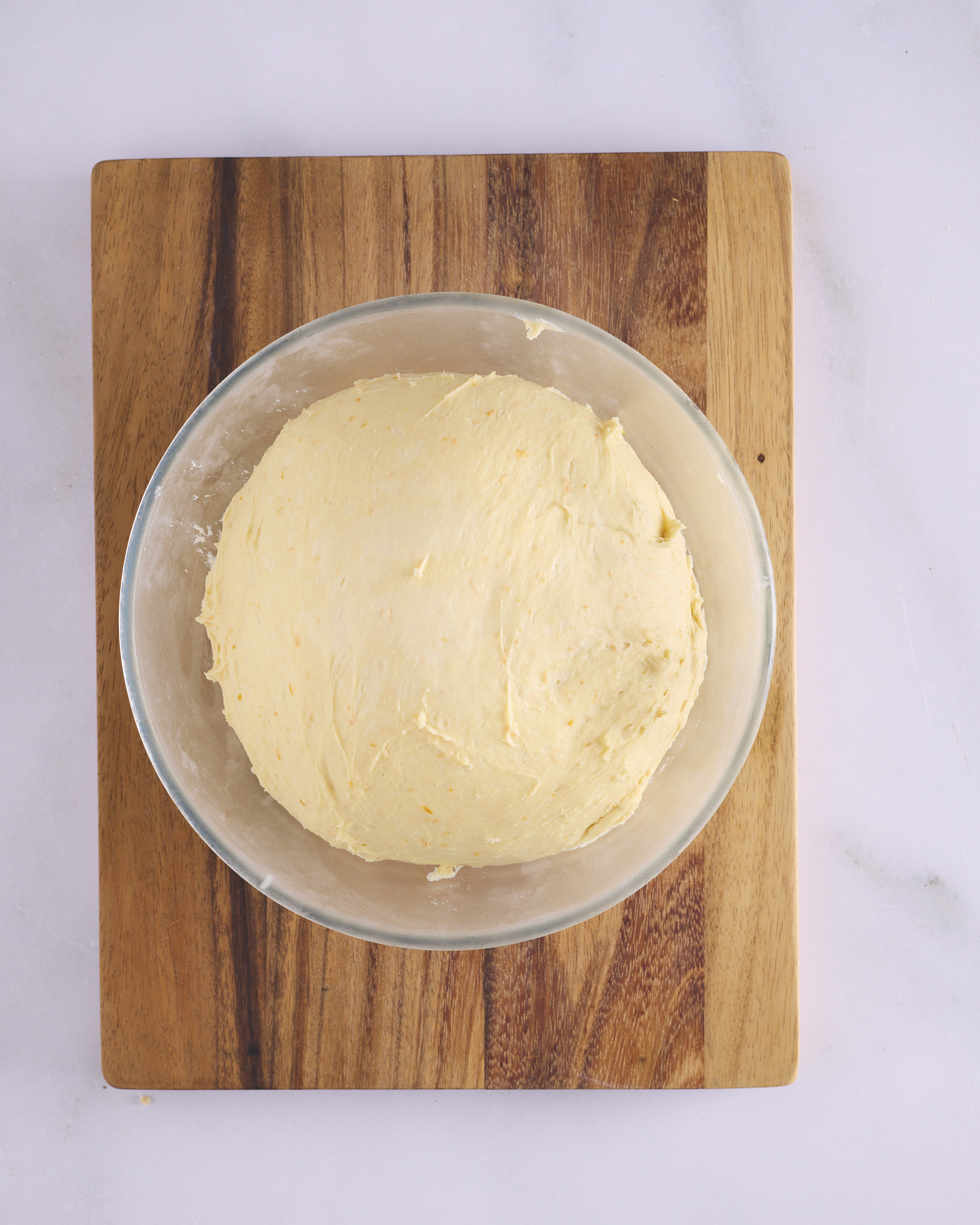 The foundation sweet dough risen in a glass bowl on a wooden board, smooth and domed, with flecks of orange zest through the surface.