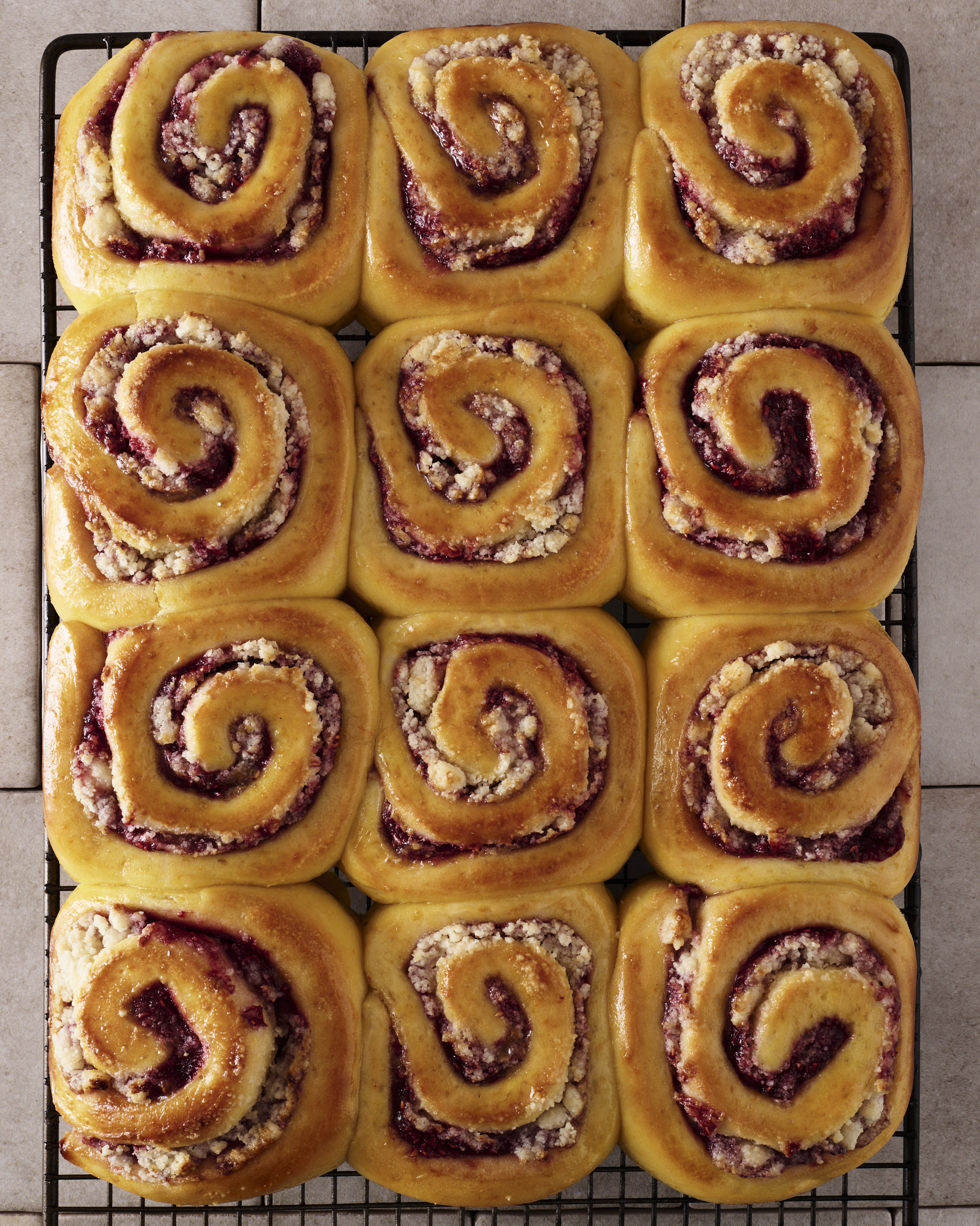 Twelve raspberry macadamia scrolls cooling on a wire rack, each swirl showing a deep purple jam through golden yeasted dough, finished with a baked crumble topping.