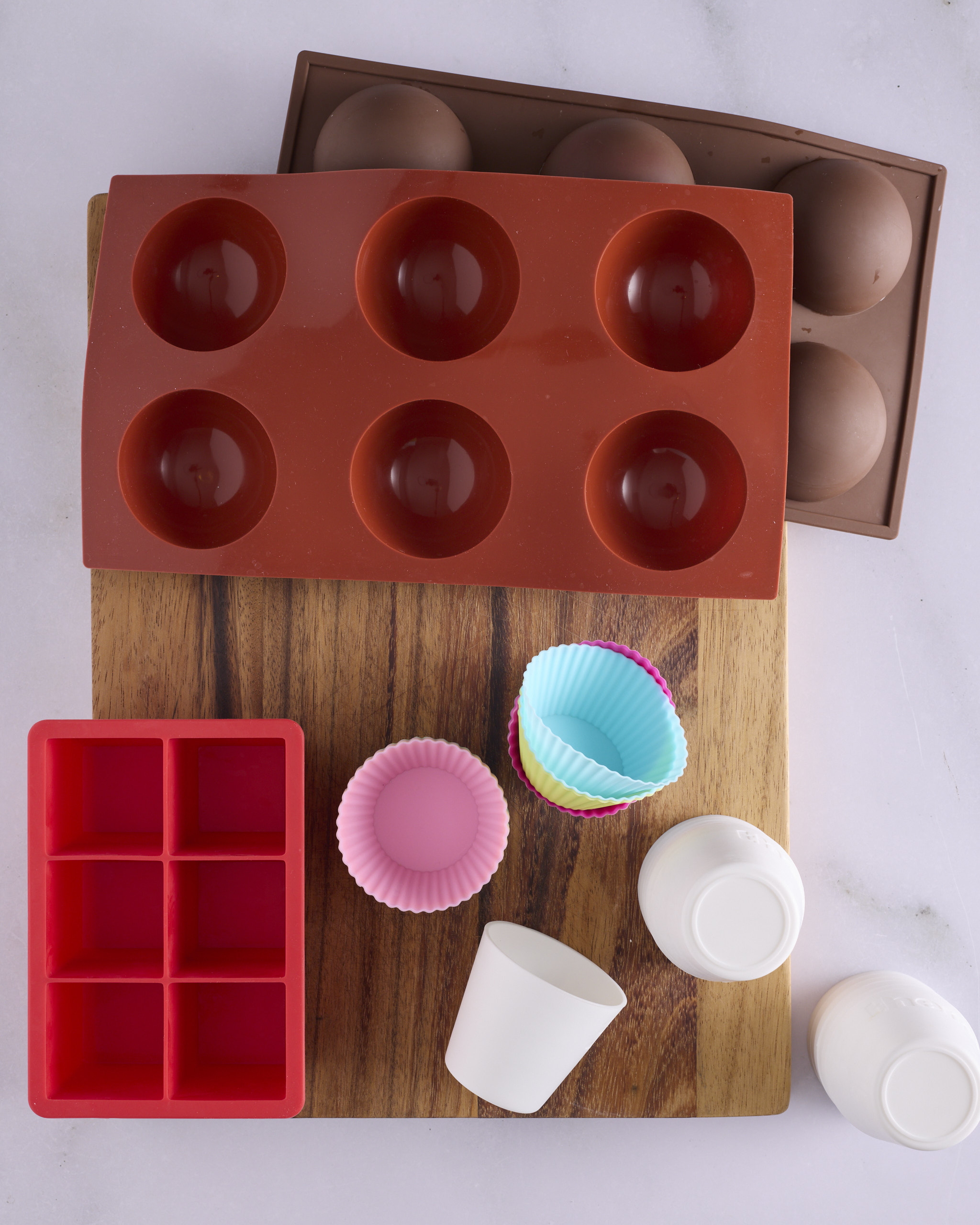 Silicone moulds for freezing the filling inserts for blueberry cheesecake kolache laid out on a wooden board. A six-hole hemisphere mould sits alongside a second hemisphere tray, a square silicone ice cube tray, patterned silicone muffin cups and white dariole moulds.