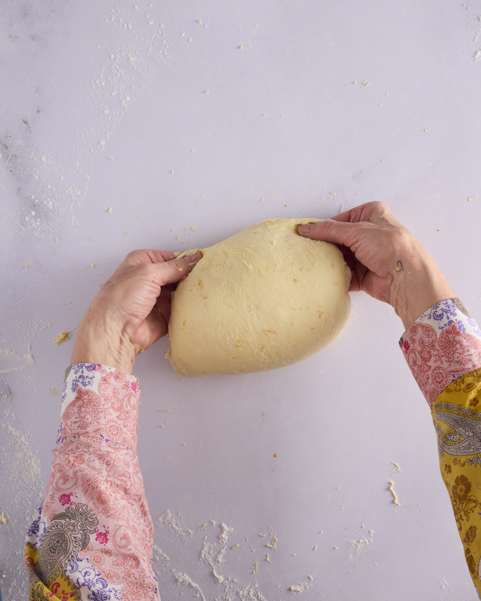 Sandra tucks the edges of the foundation sweet dough underneath the ball to build surface tension. The dough is smooth and rounded with flecks of orange zest showing through.