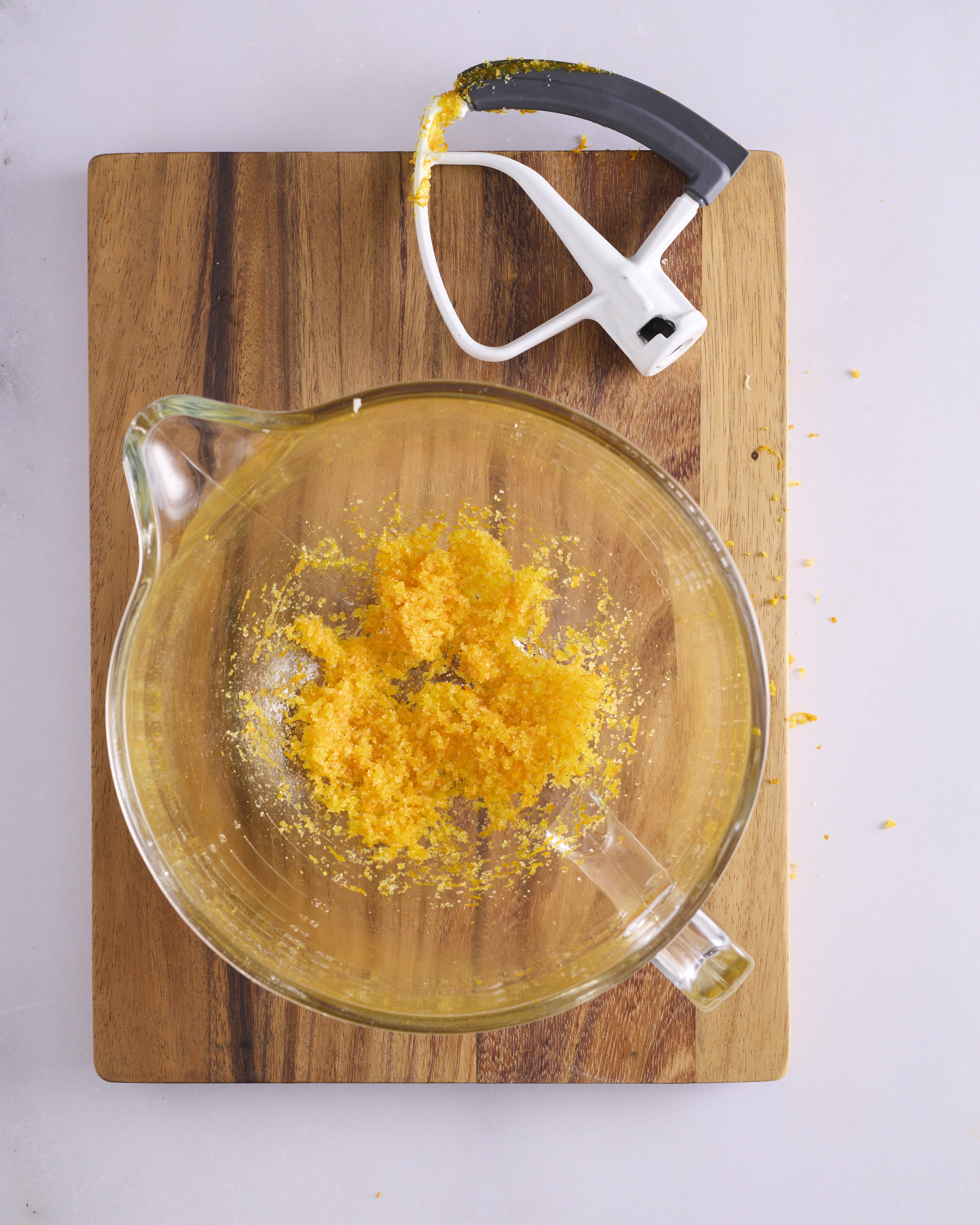 Orange zest and white sugar rubbed together in a glass stand mixer bowl for a foundation sweet dough recipe. The mixture is damp and deep orange, with the paddle attachment resting on the board beside it.