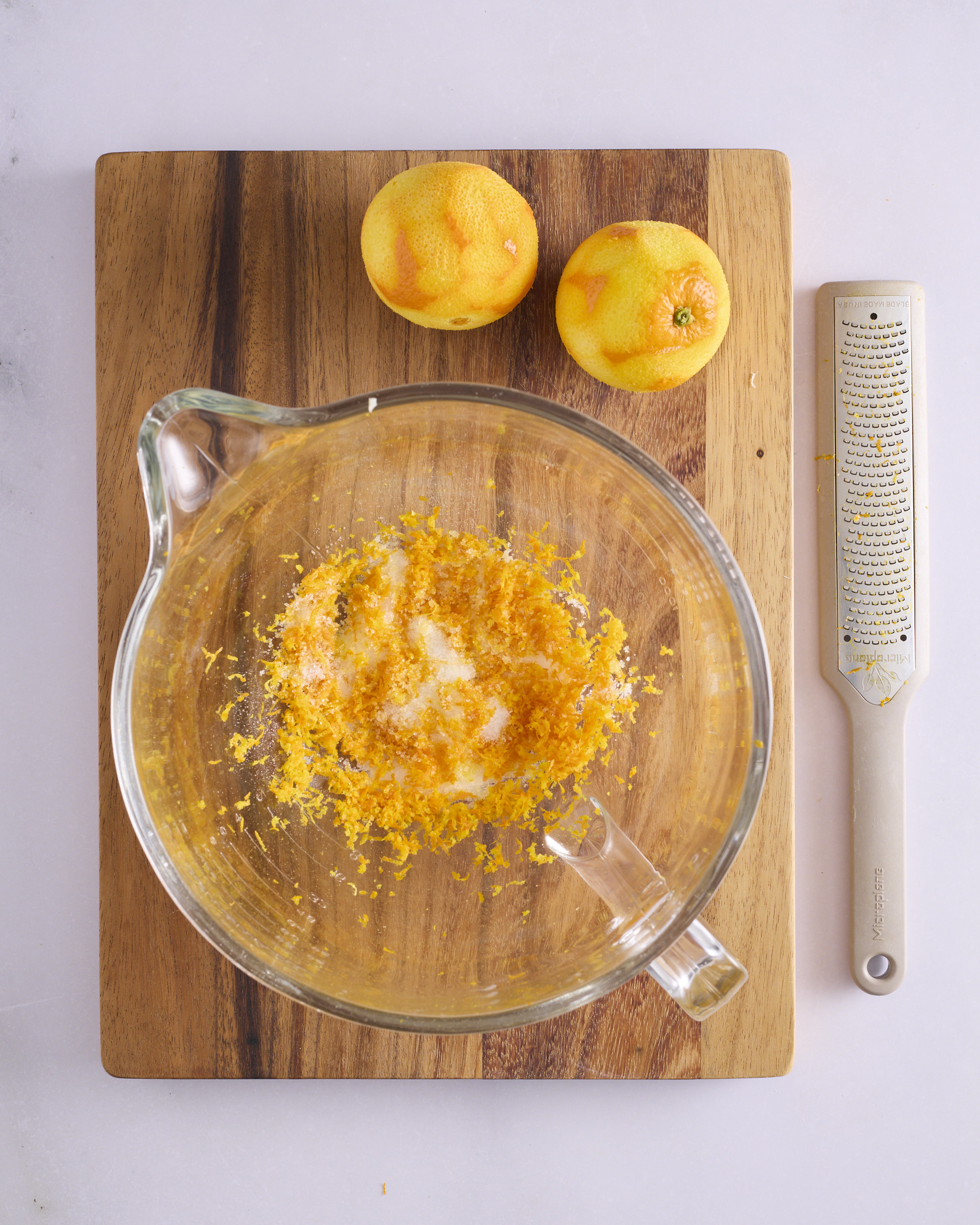 Two zested navel oranges sit on a wooden board beside a Microplane, with orange zest and white sugar in a glass stand mixer bowl ready for the foundation sweet dough.