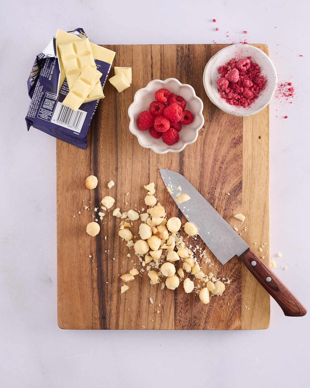 White chocolate, a bowl of fresh raspberries, a bowl of crumbled freeze dried raspberries and a pile of chopped macadamia nuts with a knife, arranged on a wooden board ready to decorate the scrolls.