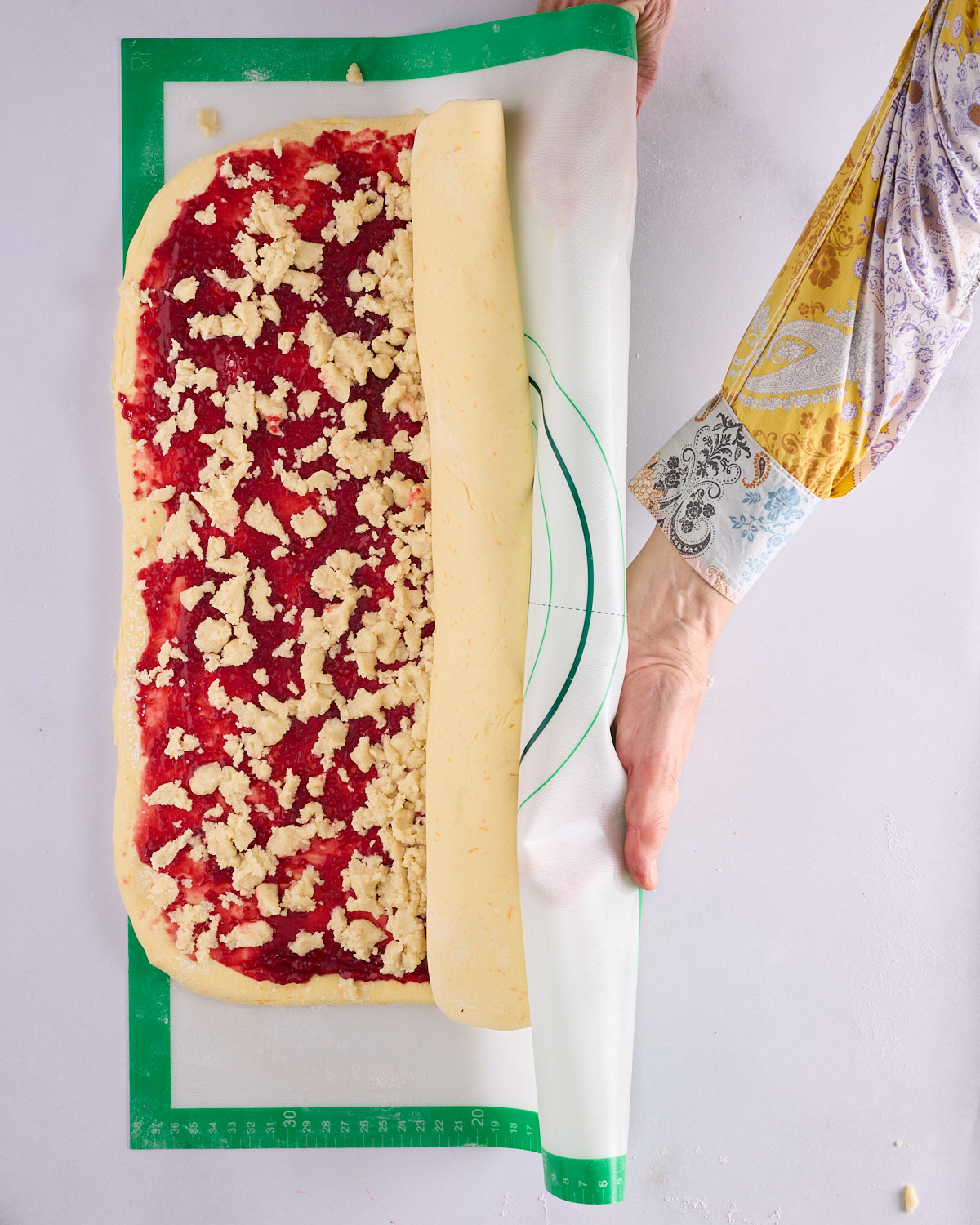 Sandra using a green silicone mat to roll the filled foundation sweet dough into a log, the jam and macadamia marzipan spiralling inside, on a marble surface.