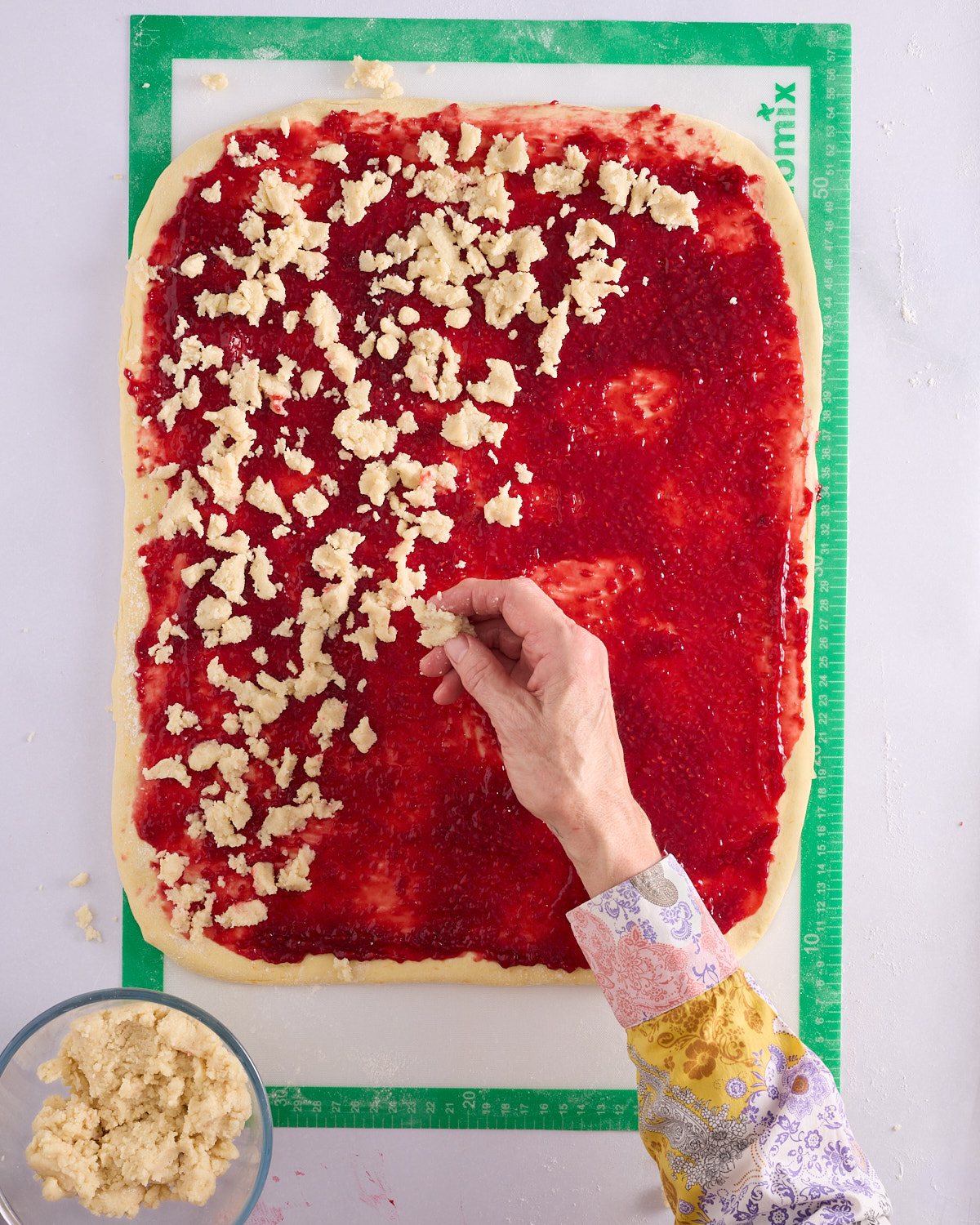 Sandra crumbling macadamia marzipan over the low sugar raspberry jam spread across the rolled foundation sweet dough on a green silicone mat, with a bowl of marzipan alongside.